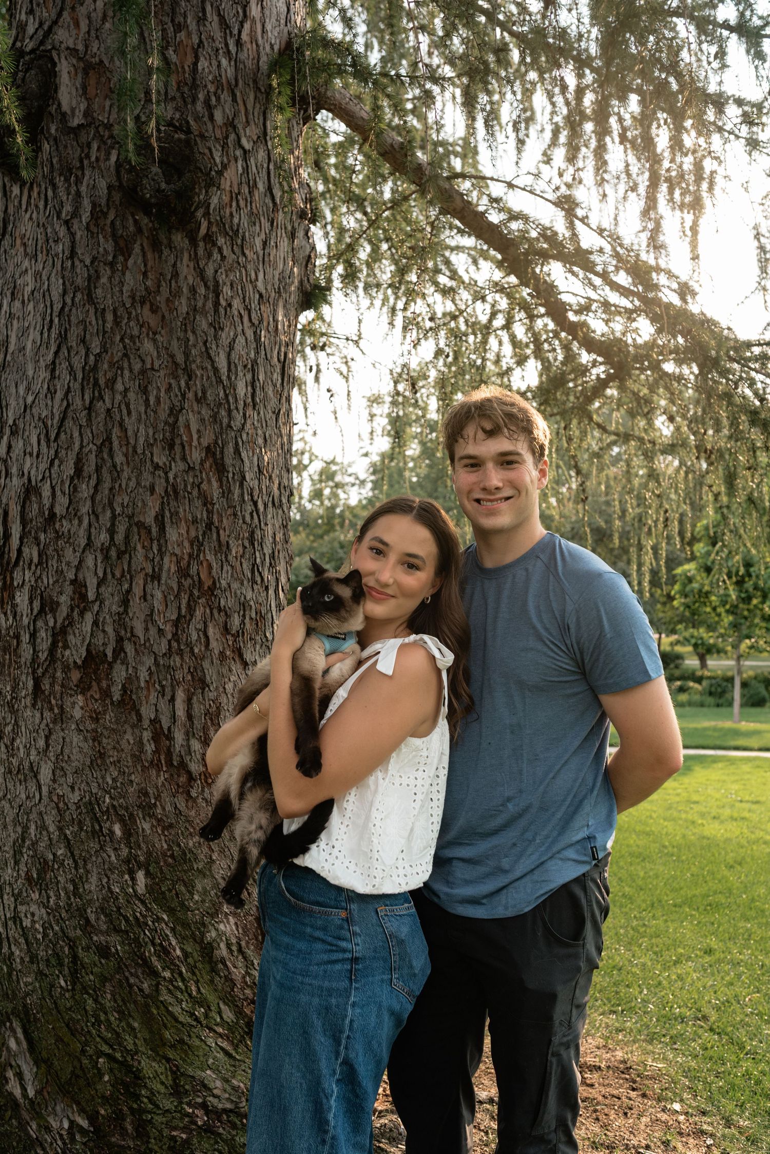 Young couple smiling outdoors by a large tree, woman holding a Siamese cat, man in blue t-shirt beside her.