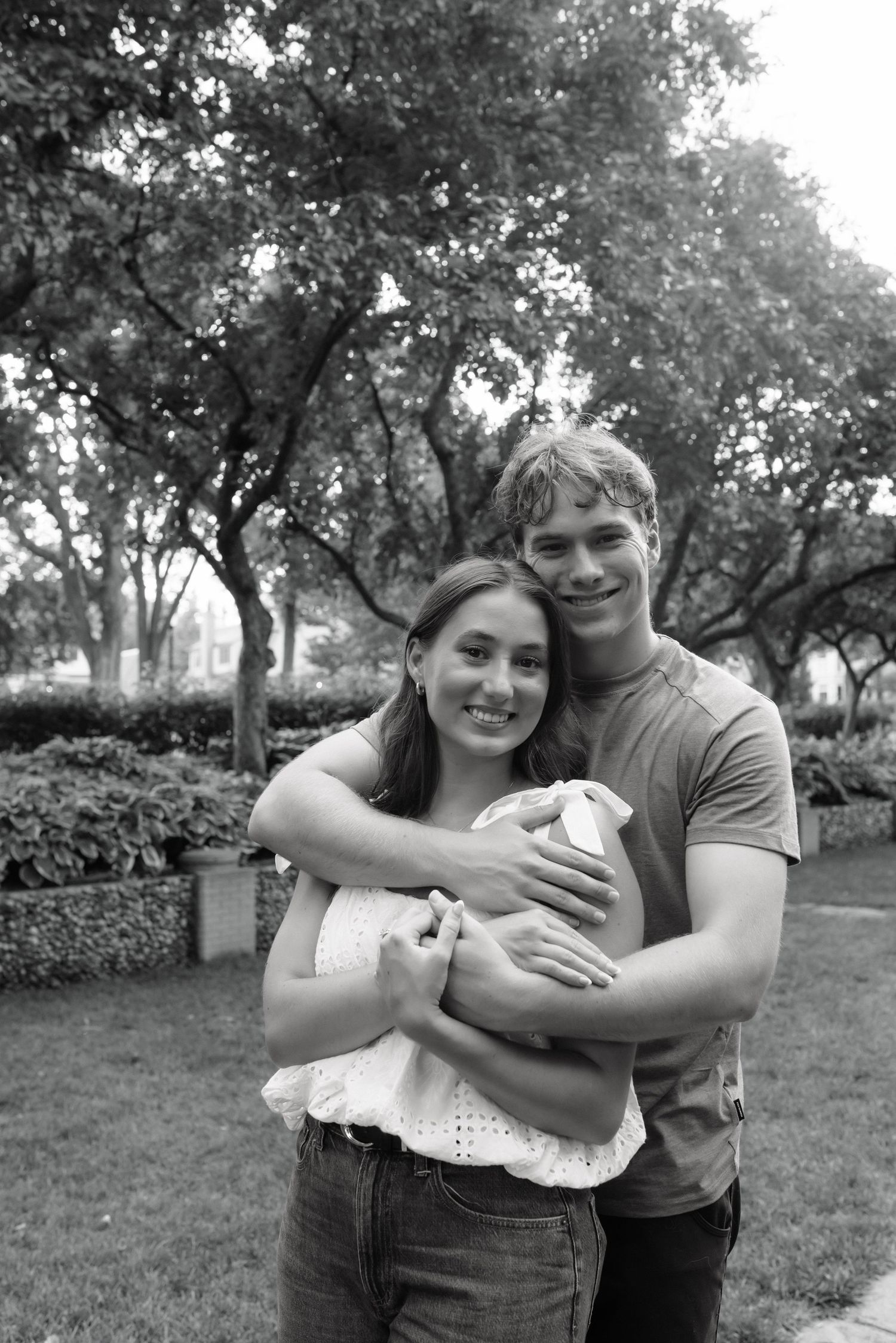 Black and white photo of a smiling young couple embracing outdoors in a park with lush trees in the background.