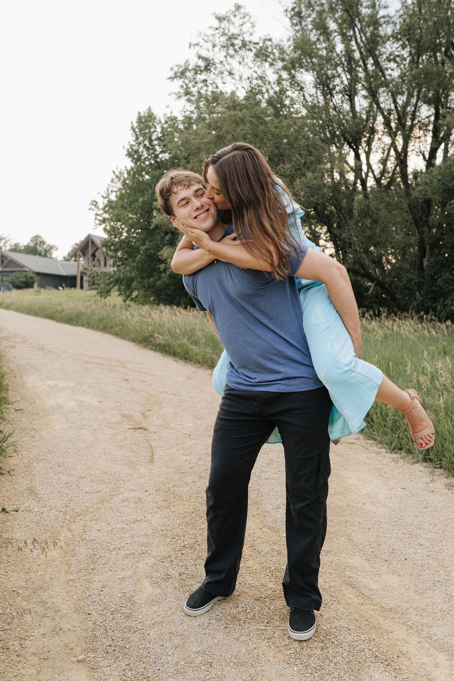 A young man giving a woman a piggyback ride on a dirt path, as she kisses his cheek outdoors.