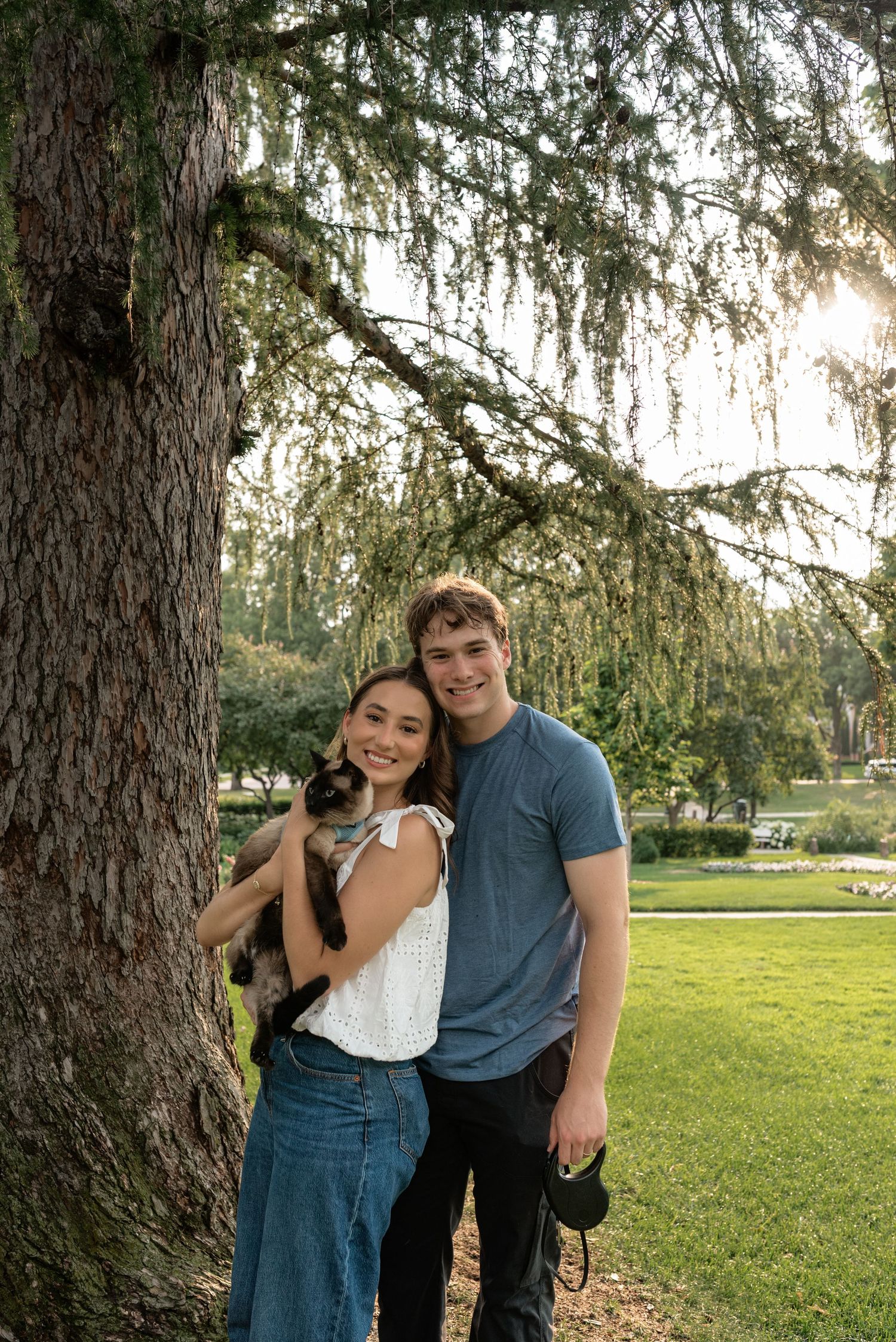 A smiling couple poses with a cat under a large tree in a sunny green park setting.