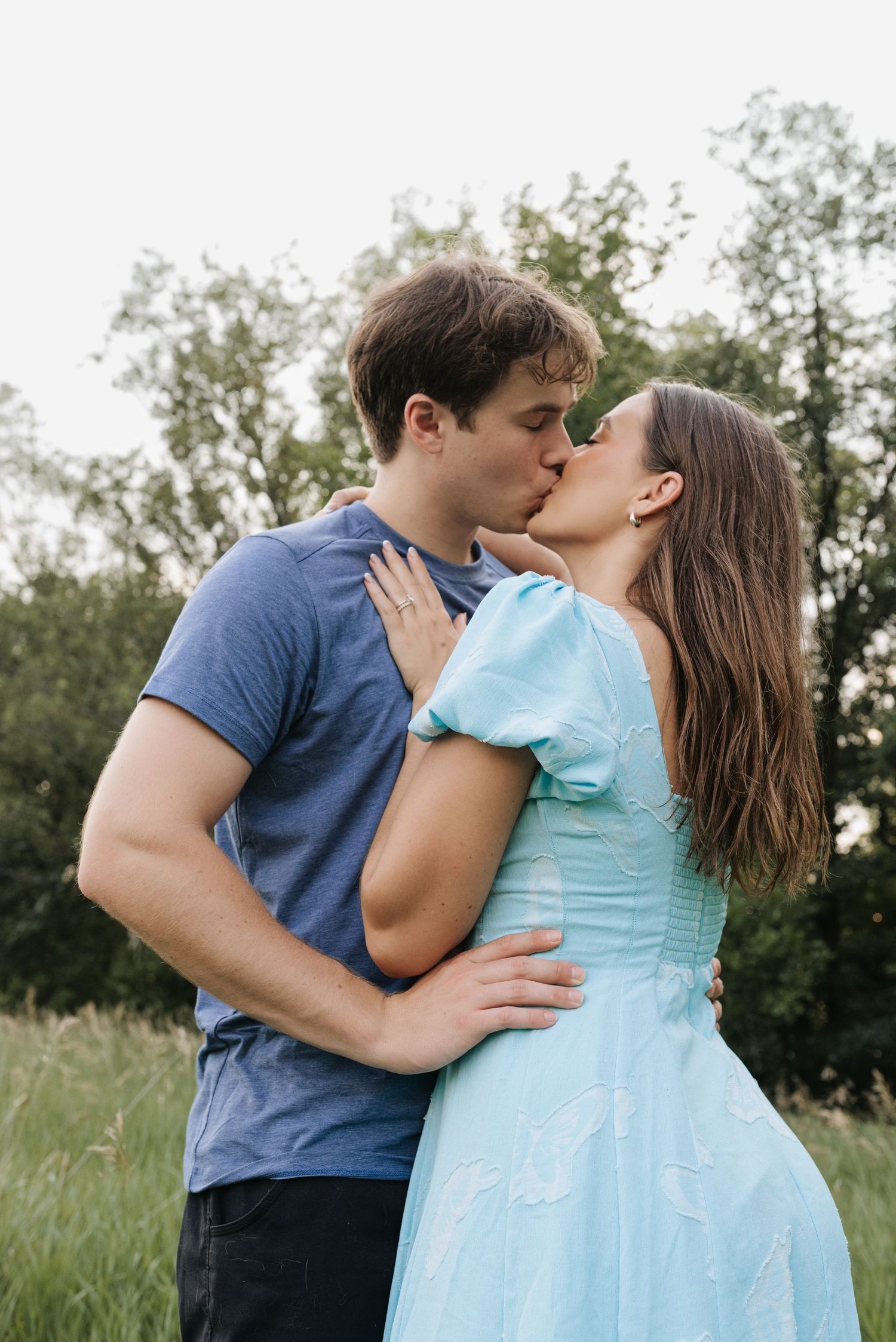 A couple shares a romantic kiss outdoors in a green field, woman in light blue dress, man in blue t-shirt.