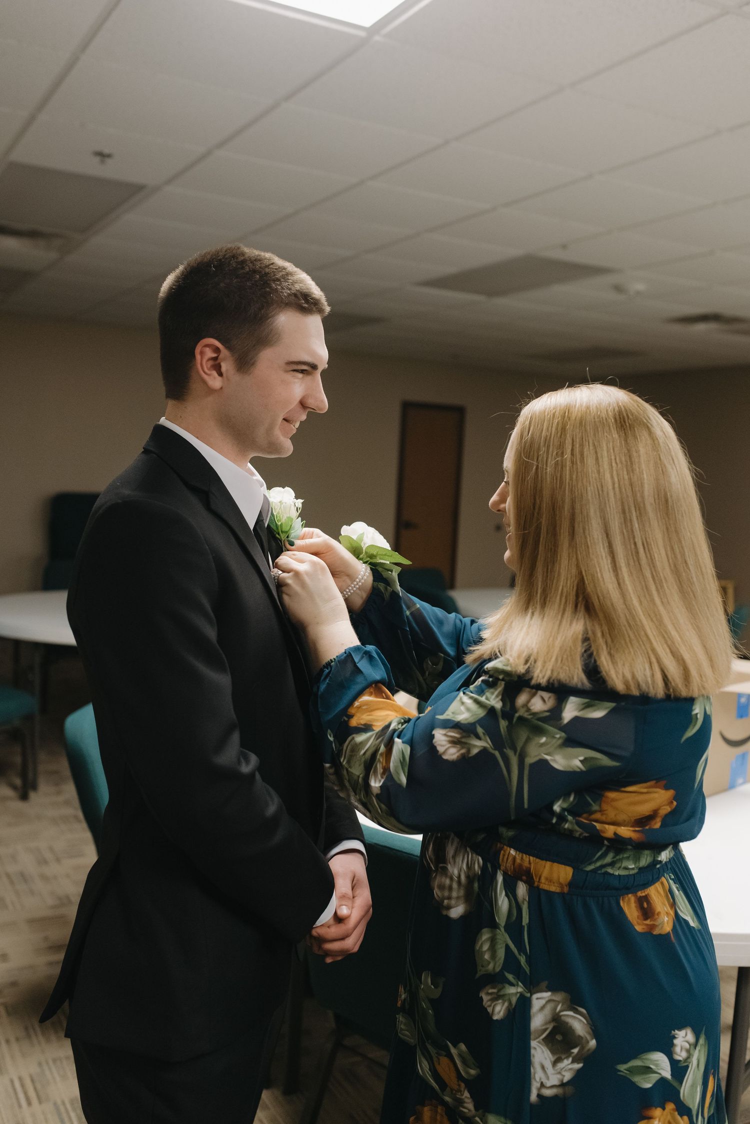 A woman in a floral dress pins a white boutonniere on a groom in a black suit before a wedding ceremony.