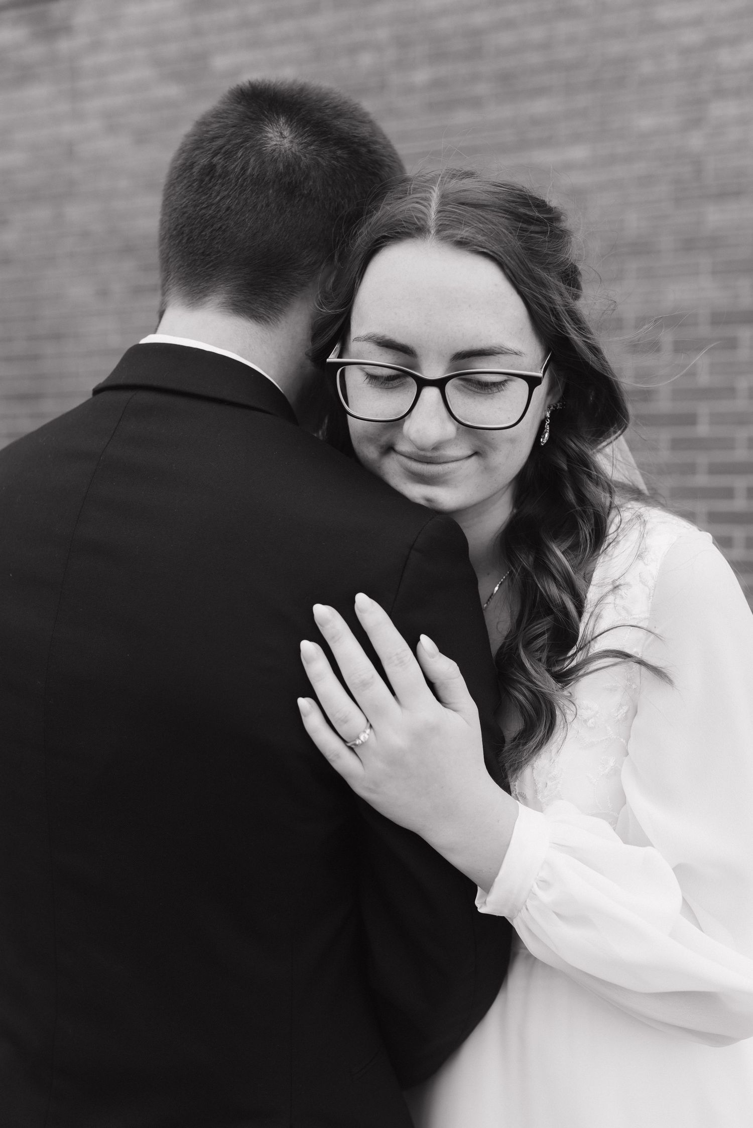 Black and white photo of bride in glasses embracing groom tenderly, showcasing her wedding ring.
