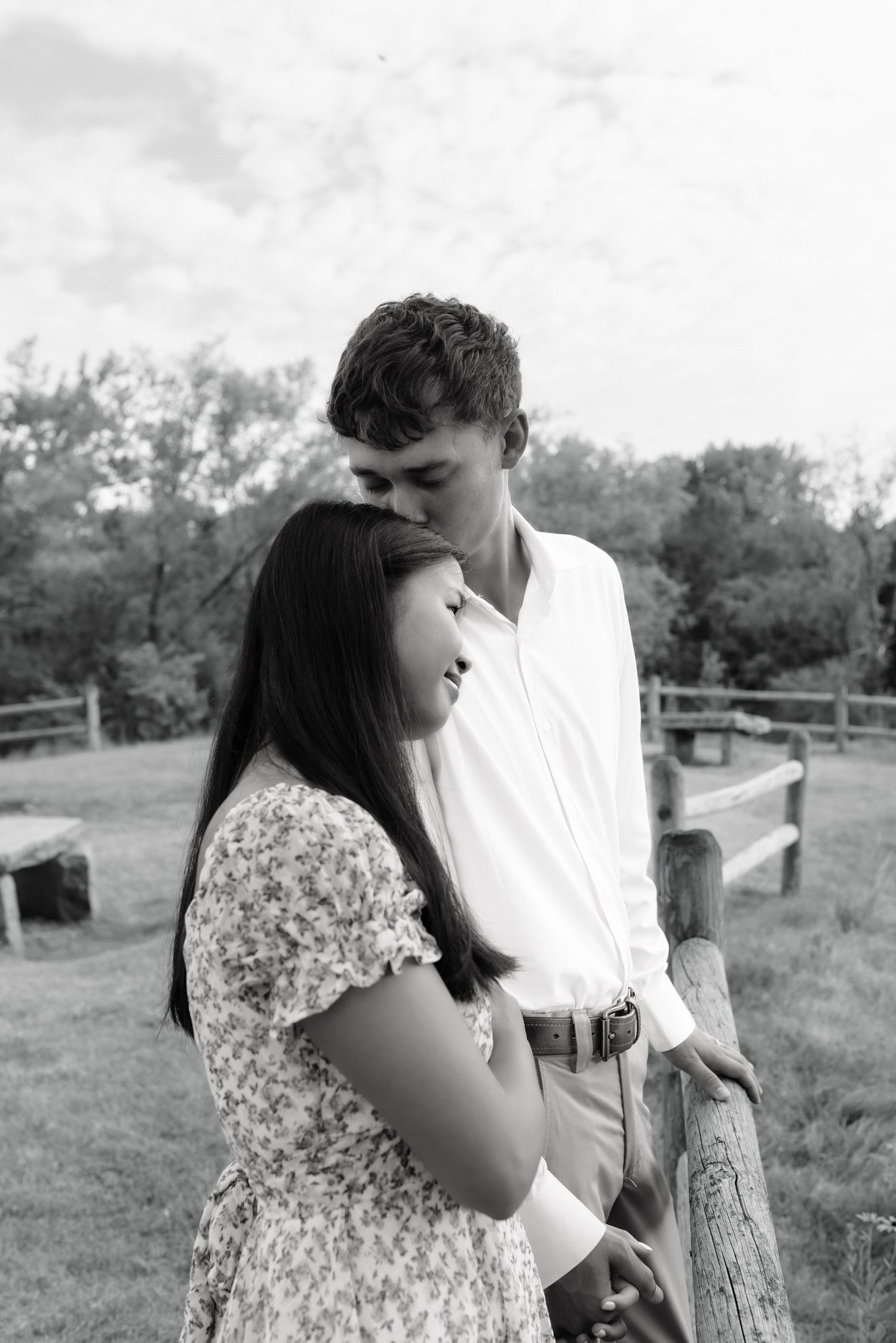 Black and white photo of couple standing by wooden fence, man kissing womans head outdoors.