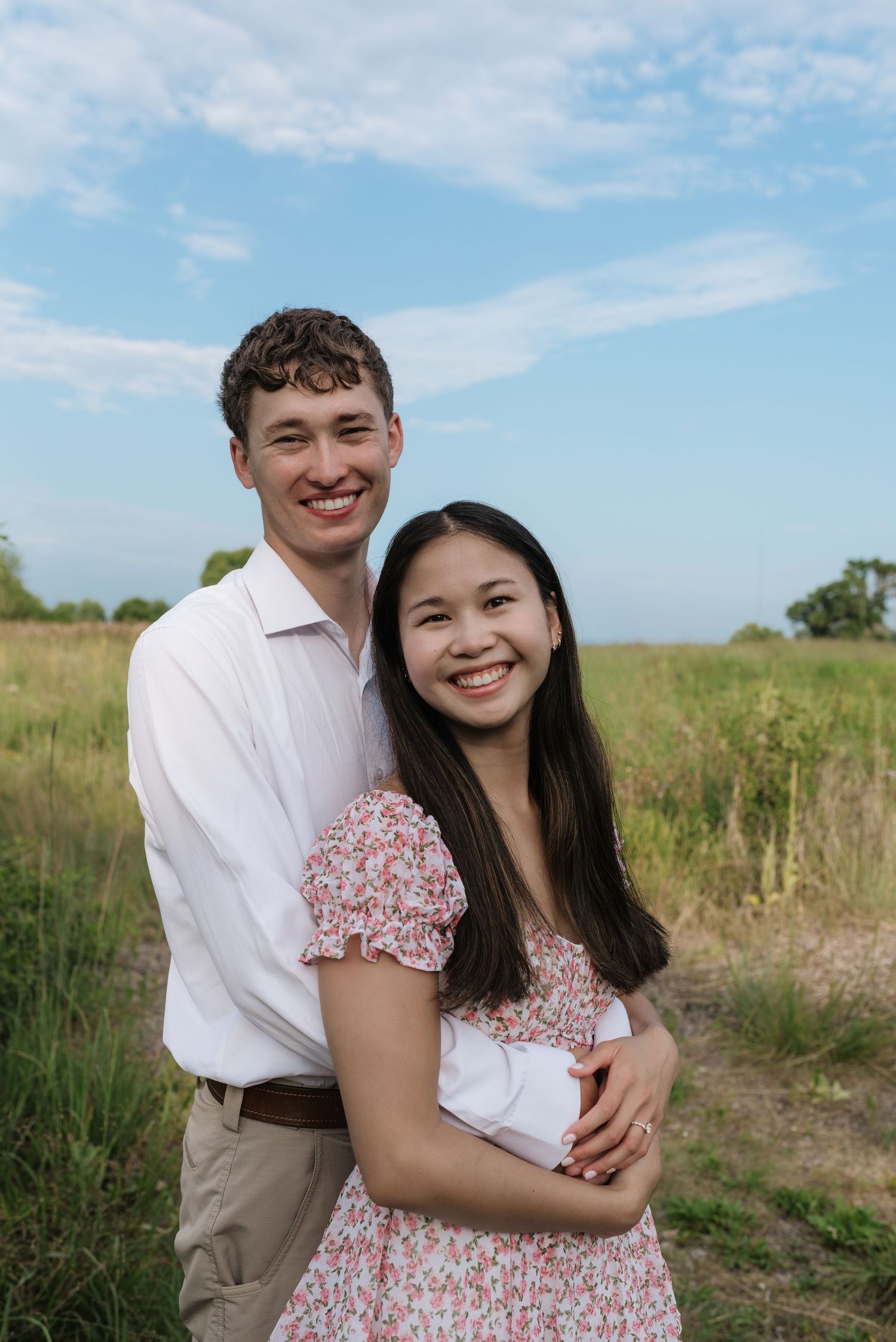 Smiling couple embracing in an open grassy field under a bright blue sky during engagement photos.