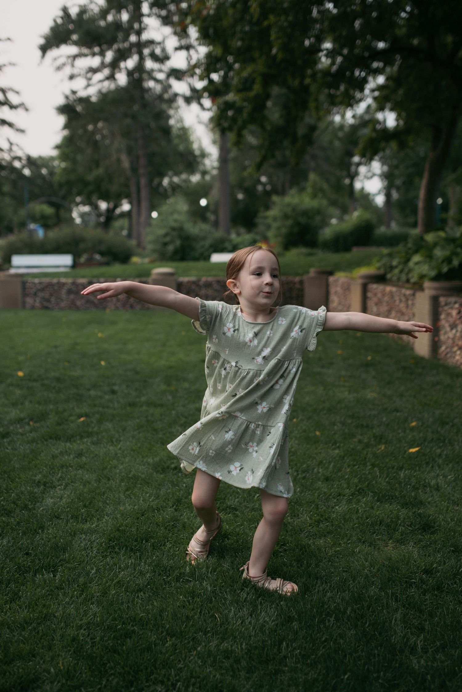 A young red-haired girl in a mint green floral dress stands with arms outstretched on a lush green lawn.