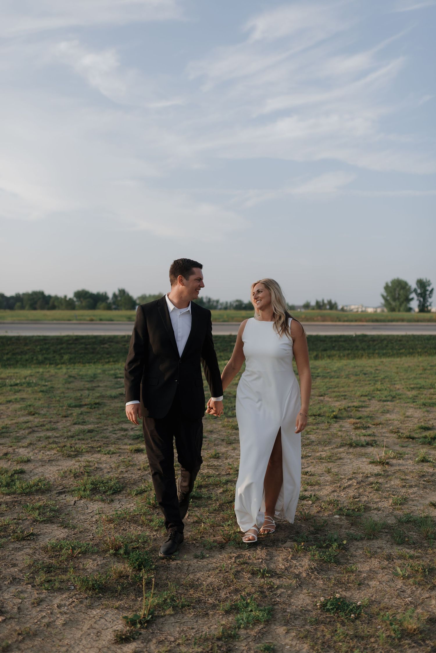 Bride in white gown and groom in black suit walking hand in hand laughing outdoors.