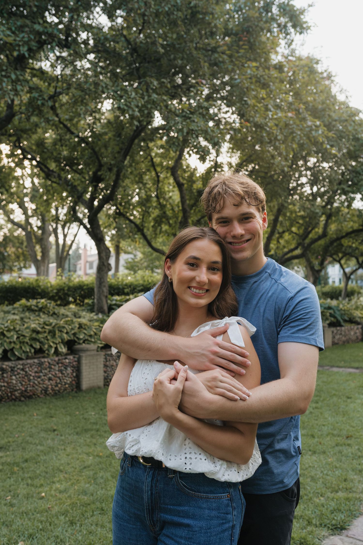 A young couple embracing and smiling in a lush green park, wearing casual blue and white outfits.