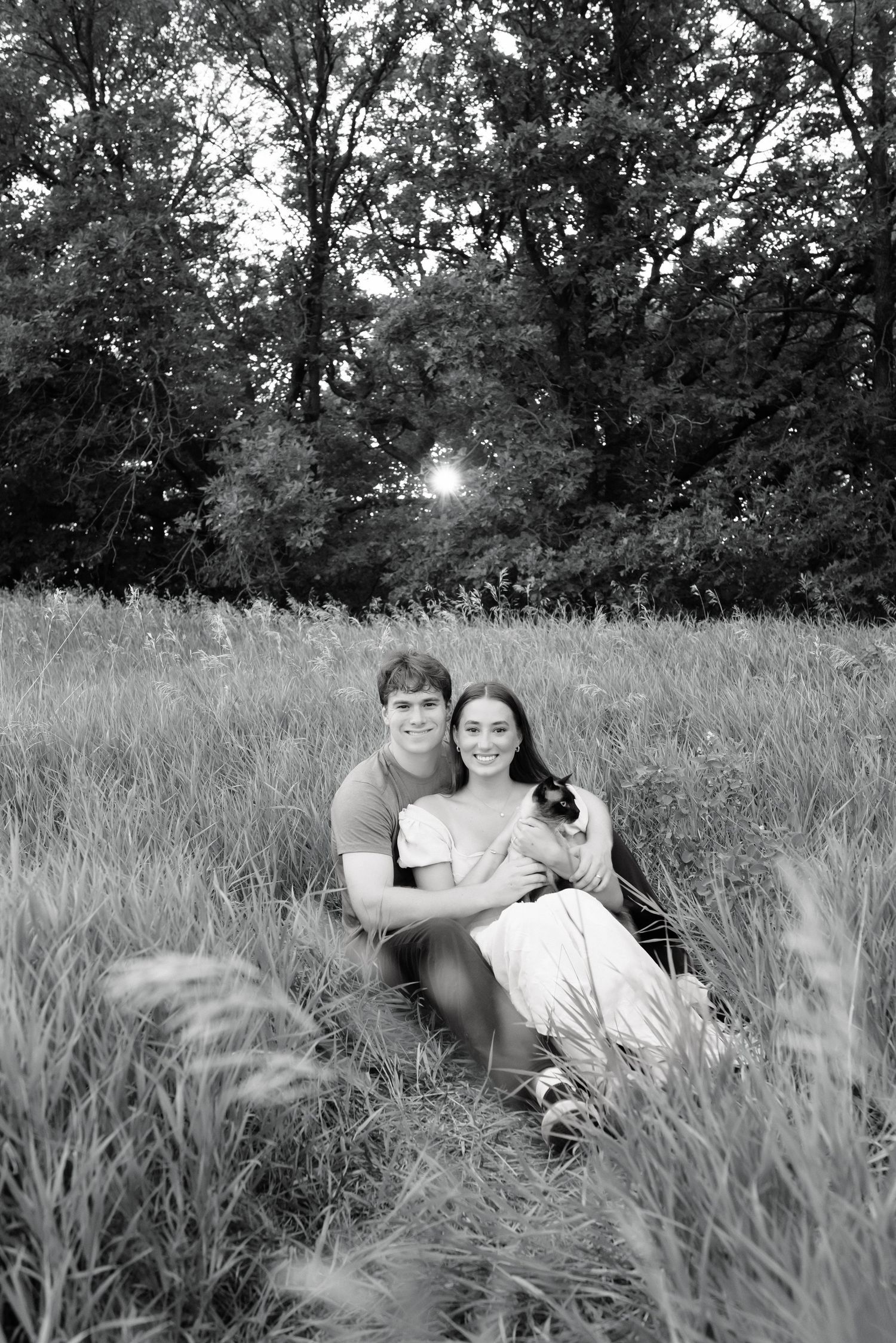 Black and white photo of a smiling couple sitting in a grassy field with trees in the background, holding a small cat.