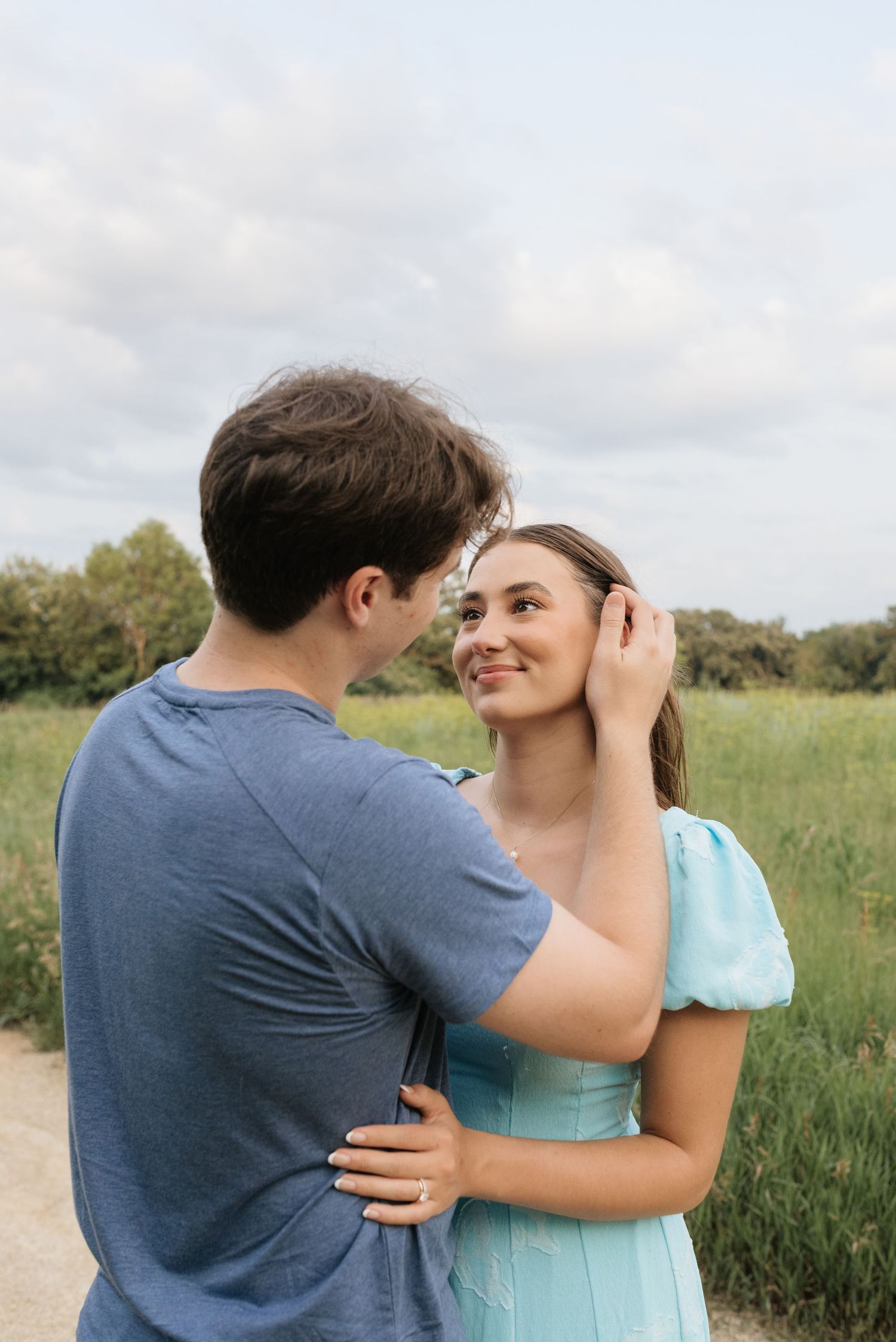 A couple shares a tender moment outdoors, man in blue tee gently touches woman's hair in a green field.