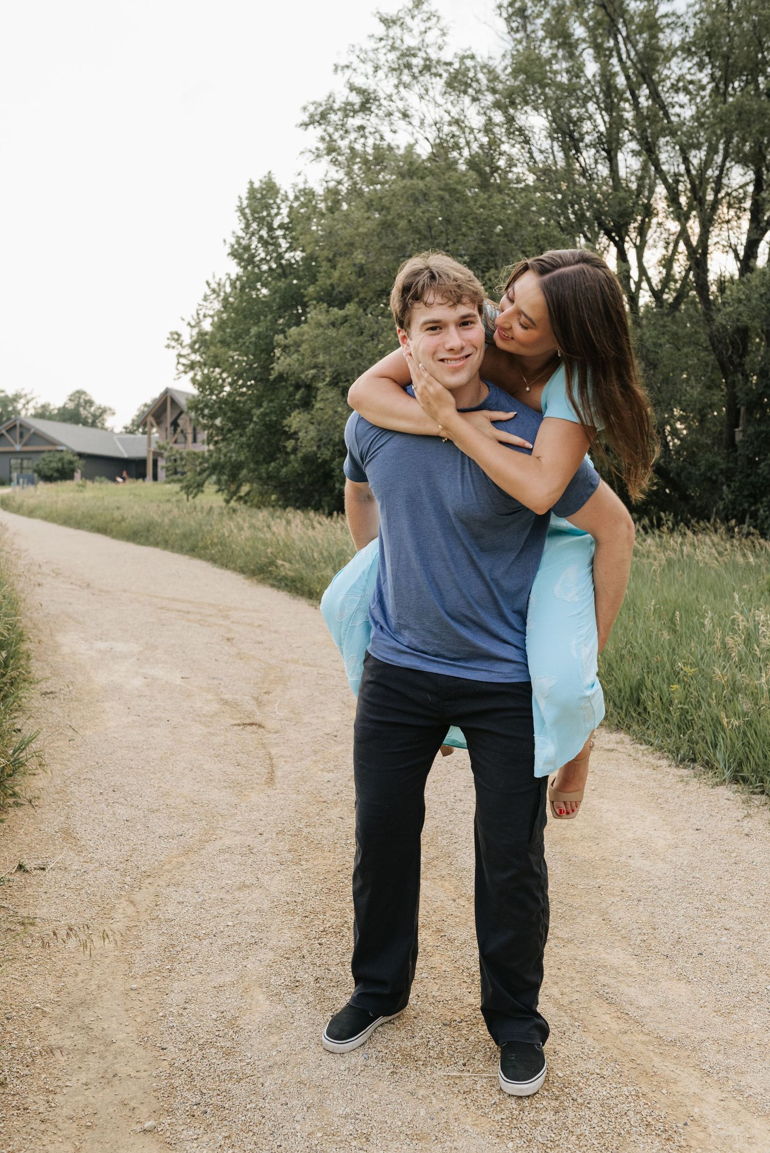 A young couple shares a piggyback ride on a rural dirt path, surrounded by lush green trees.