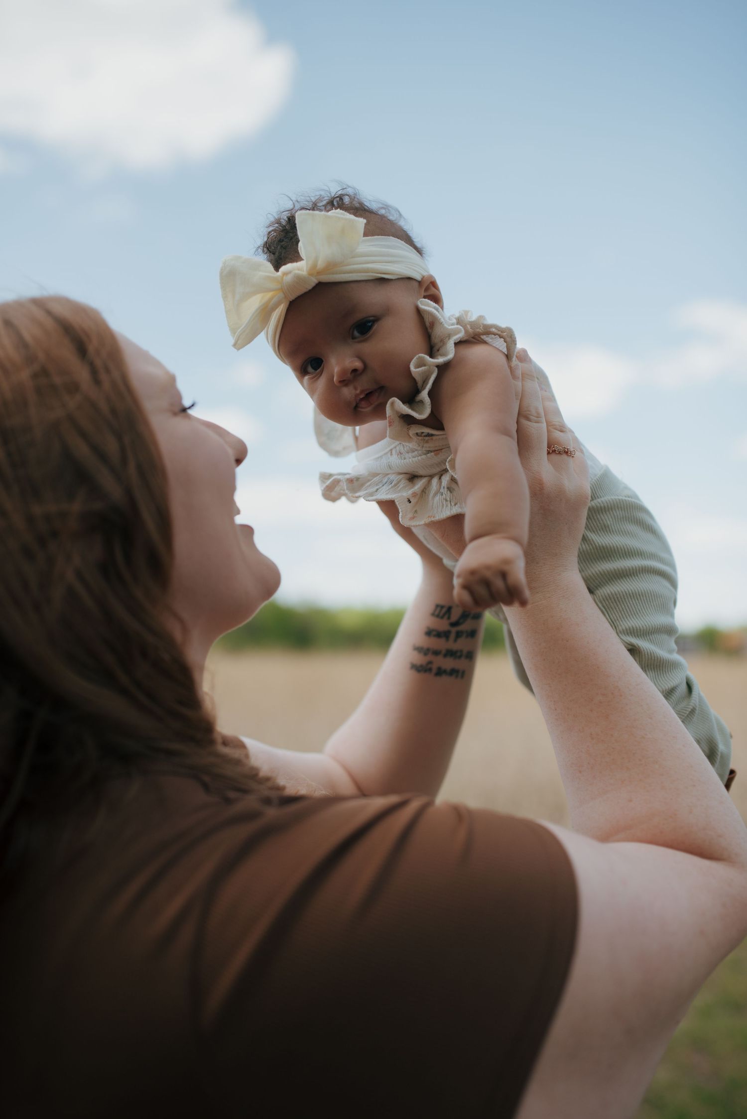 Joyful mother lifting smiling baby girl in white bow headband outdoors under blue sky.