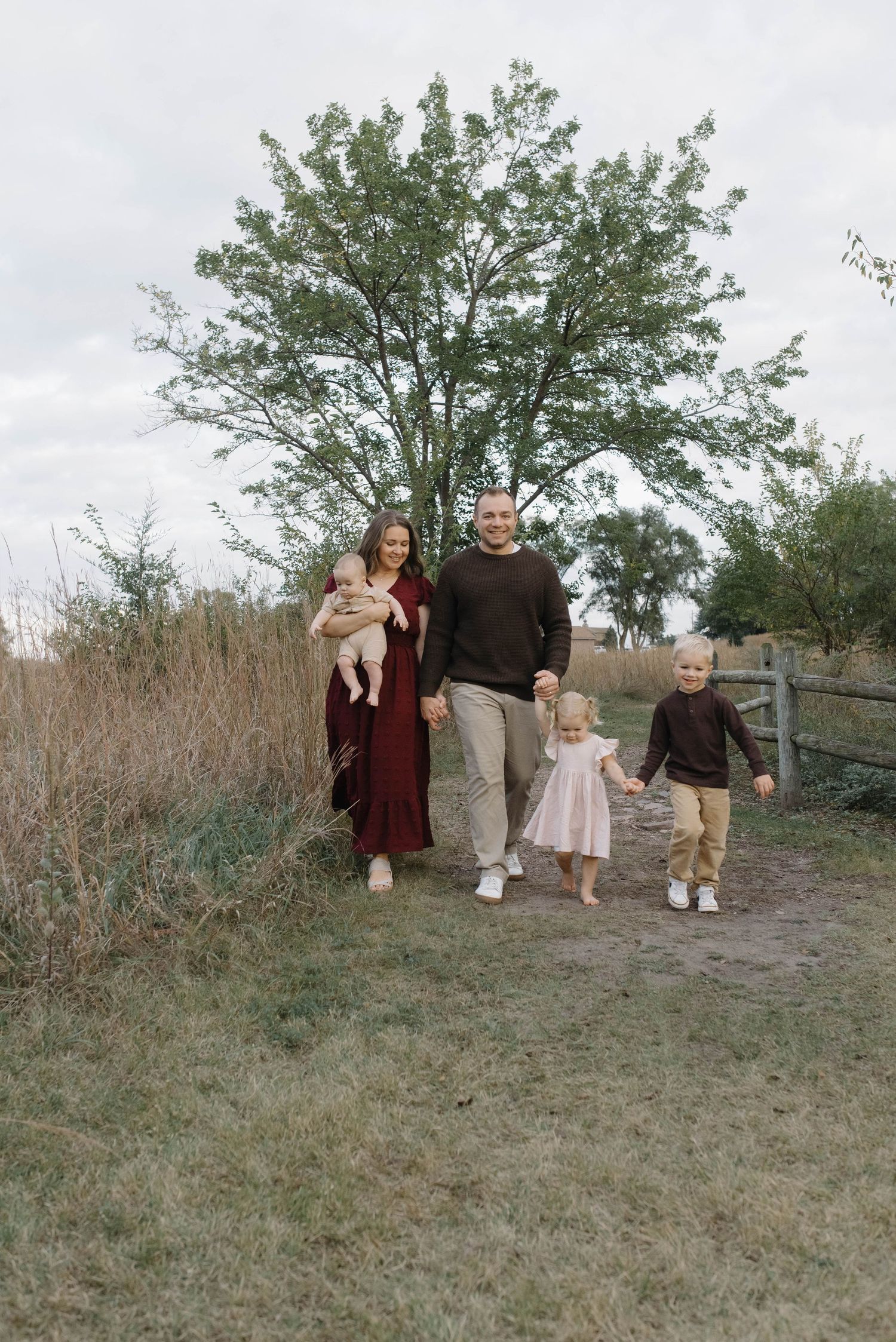 Smiling family of four holding hands while walking on a dirt path with trees in background.