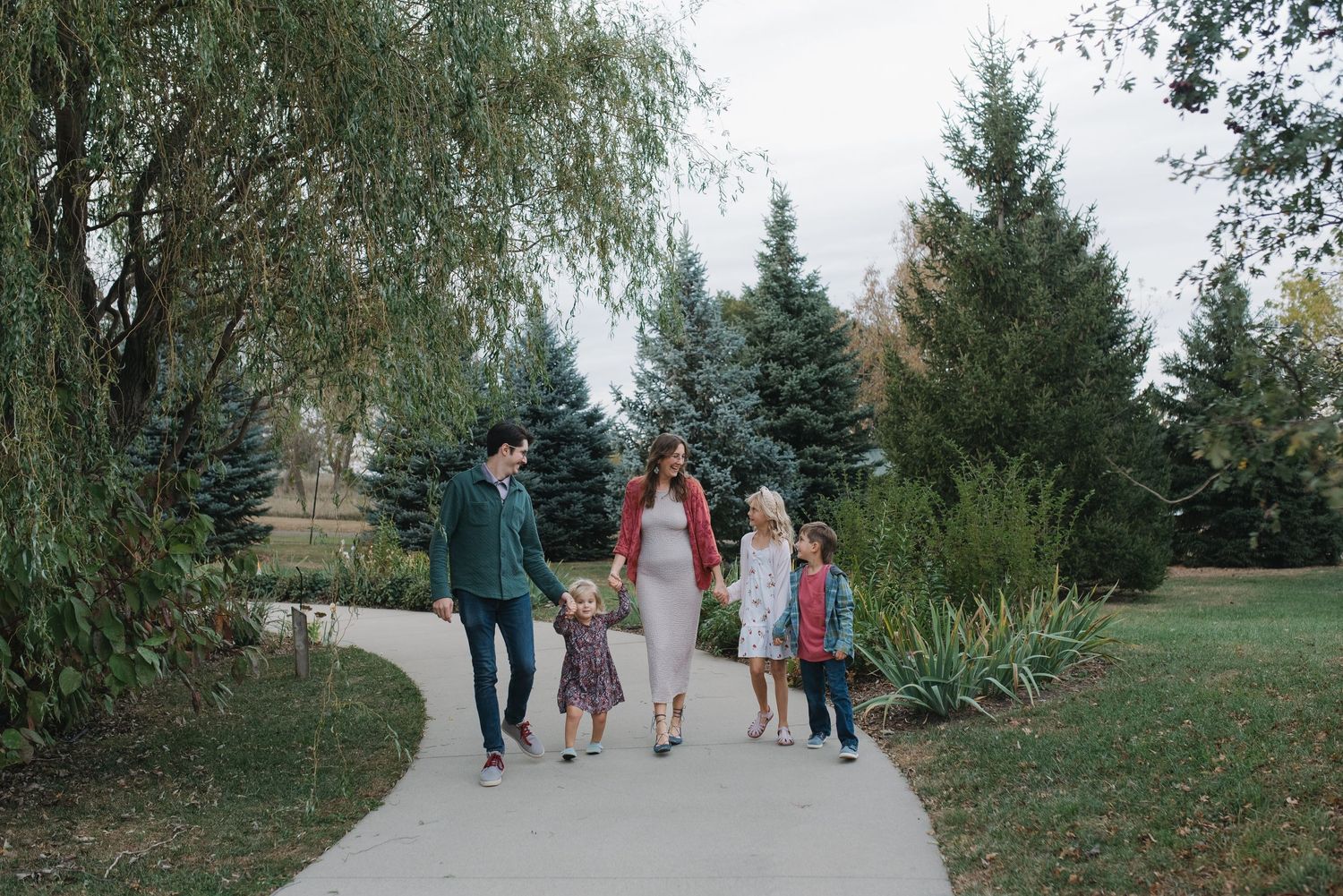 A pregnant woman and her family walk hand-in-hand along a tree-lined park path.