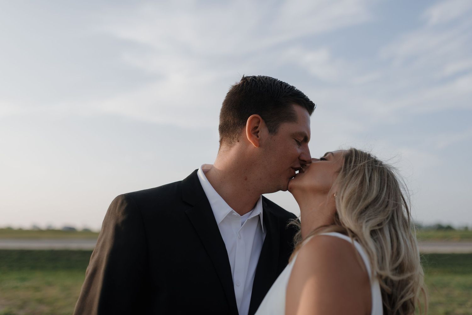 Newlyweds kissing tenderly in an open field with a soft cloudy sky backdrop.
