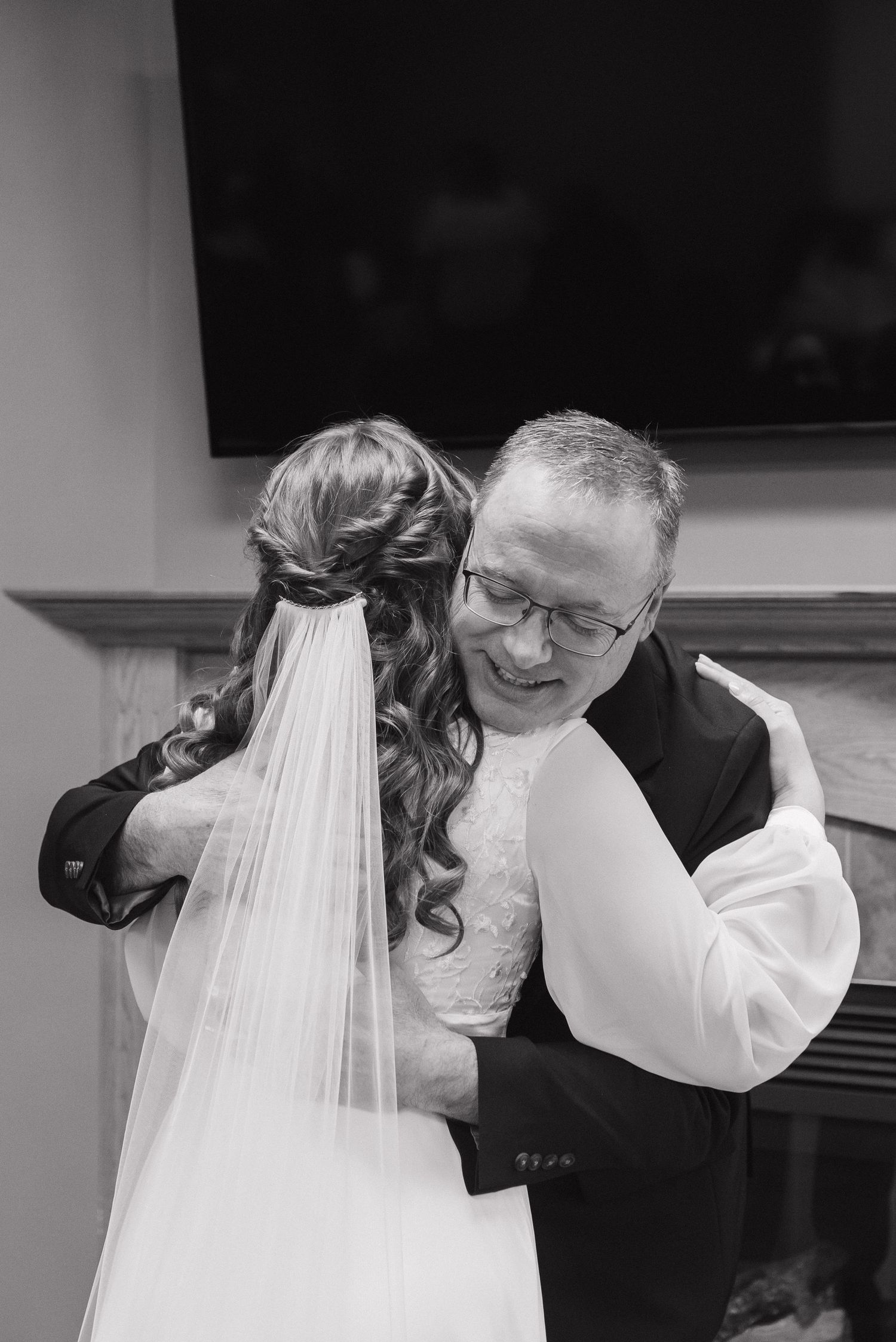Black and white photo of a bride in a veil embracing a smiling older man in glasses at a wedding.