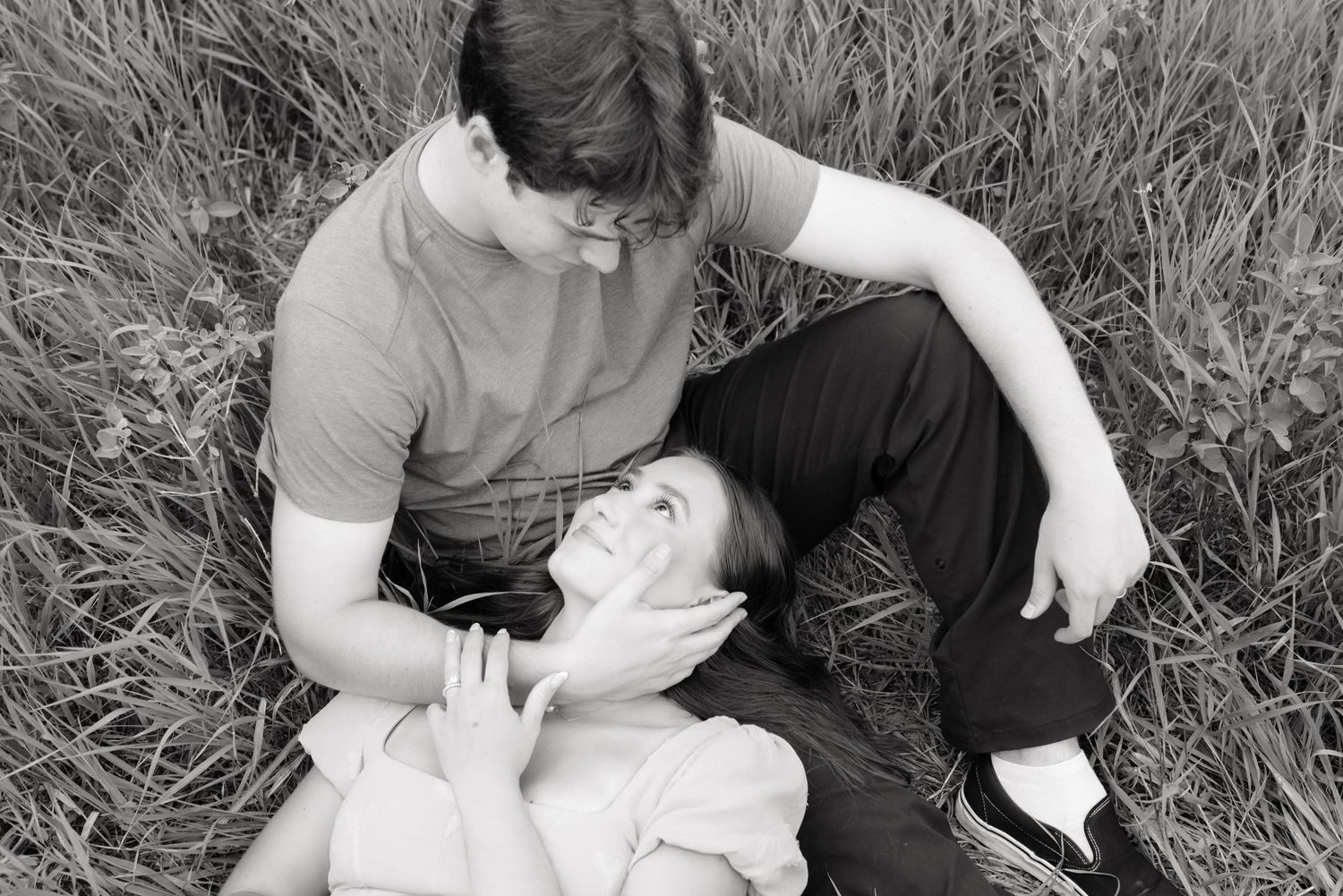 Black and white photo of a young couple sitting in tall grass, woman lying in mans lap gazing up at him.