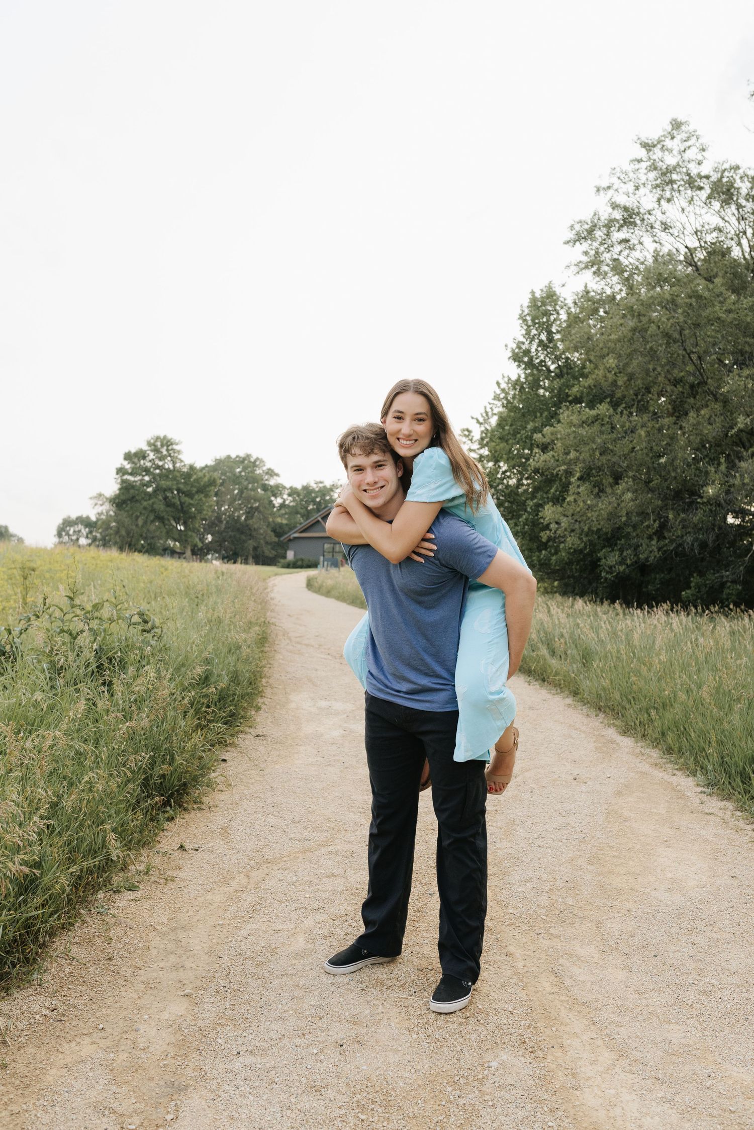 A young couple smiling on a dirt path, woman giving piggyback ride, surrounded by green fields and trees.