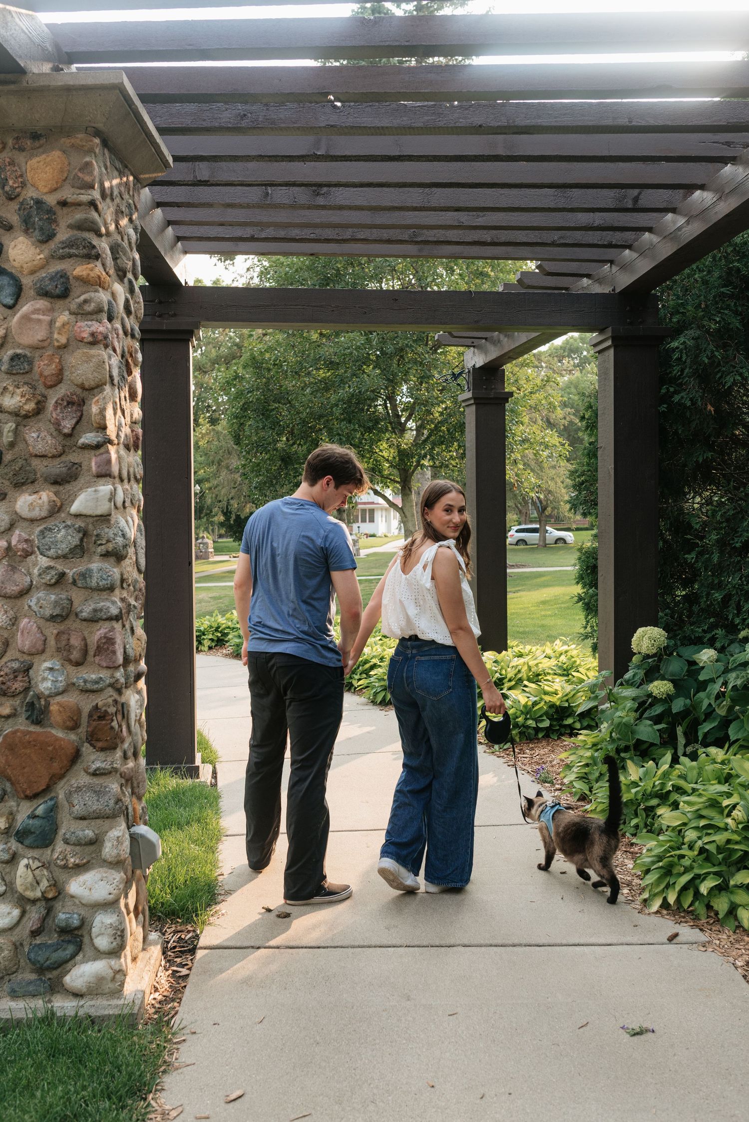 Couple holding hands walking their cat on a leash under a stone pergola surrounded by lush green gardens.
