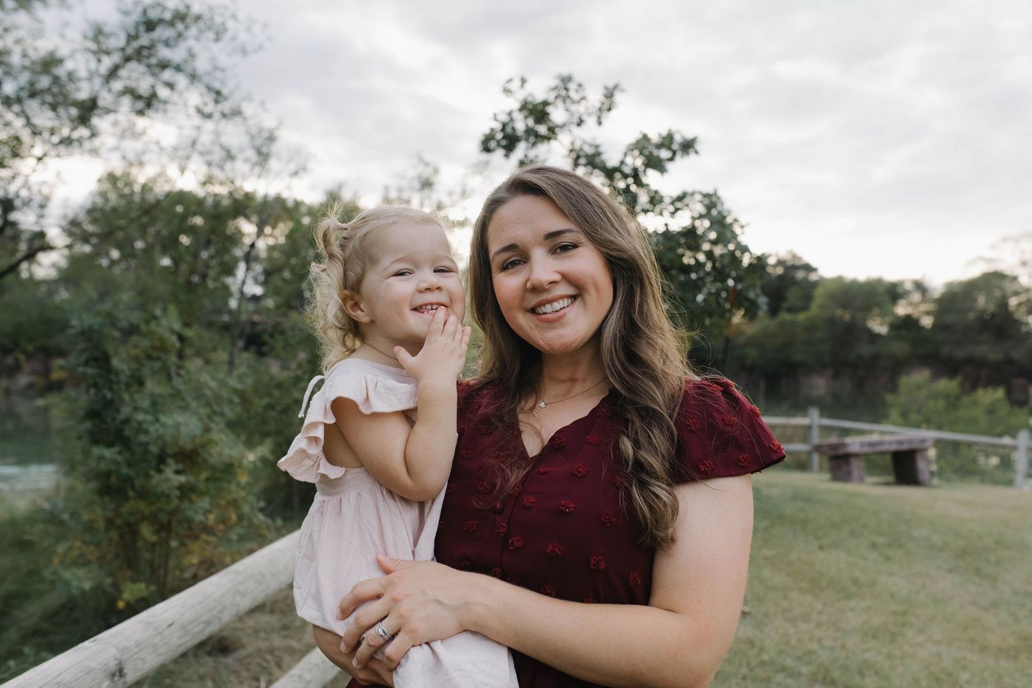 Smiling mother holding young blonde daughter outdoors near wooden fence with green trees.