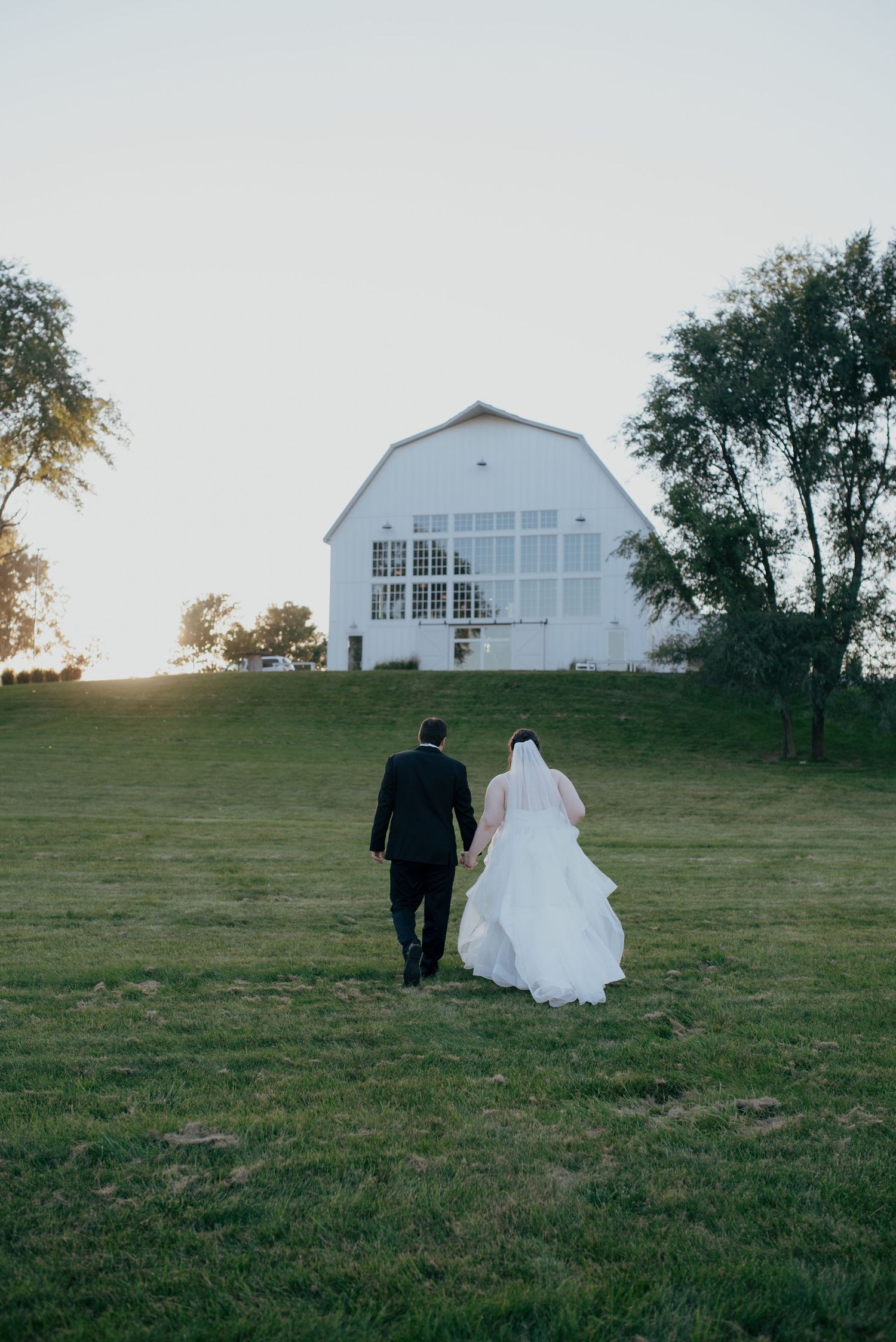 Bride and groom walking hand in hand toward a white barn venue on a lush green hill at sunset.