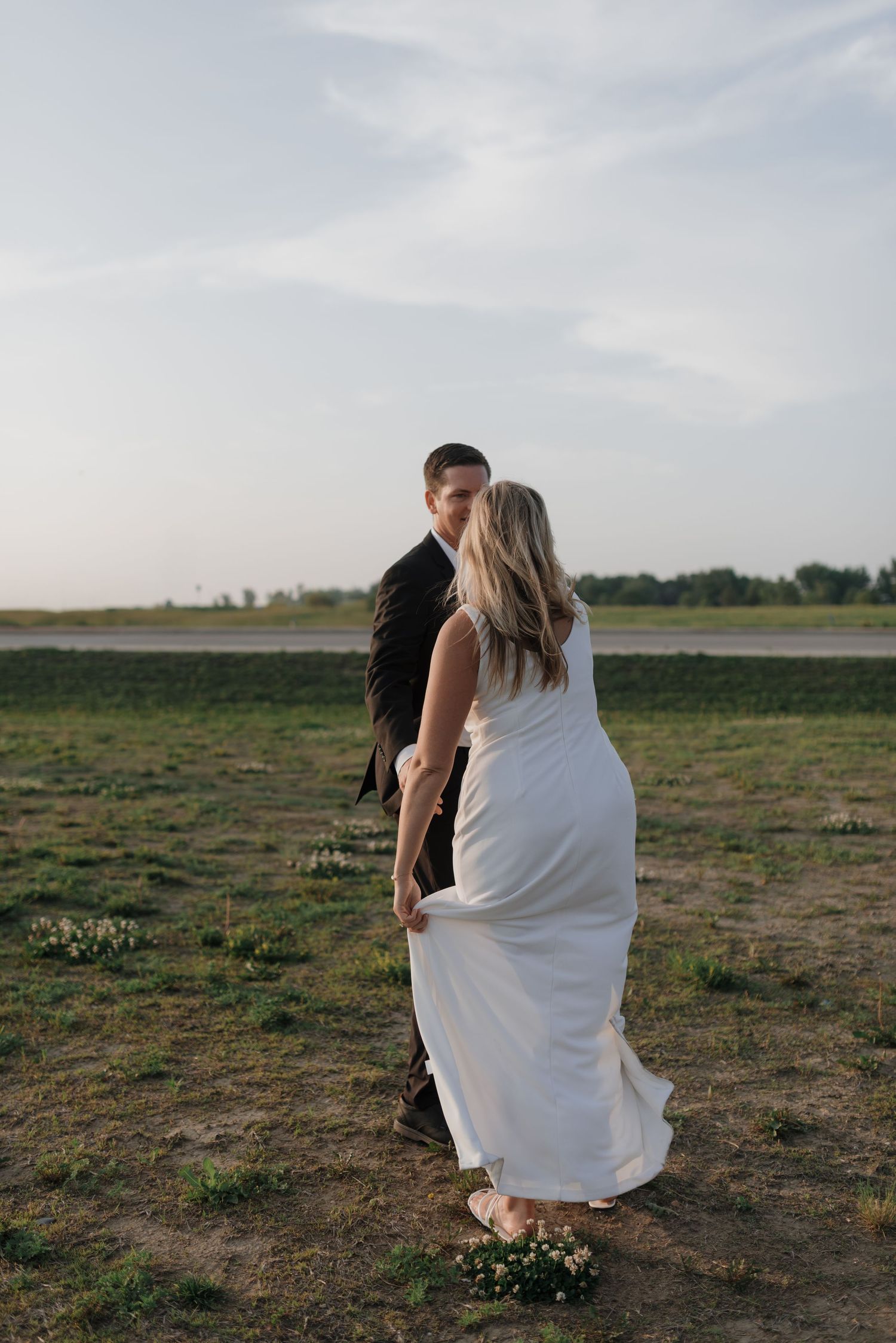 Newlywed couple walks hand in hand through open field during sunset wedding photography.