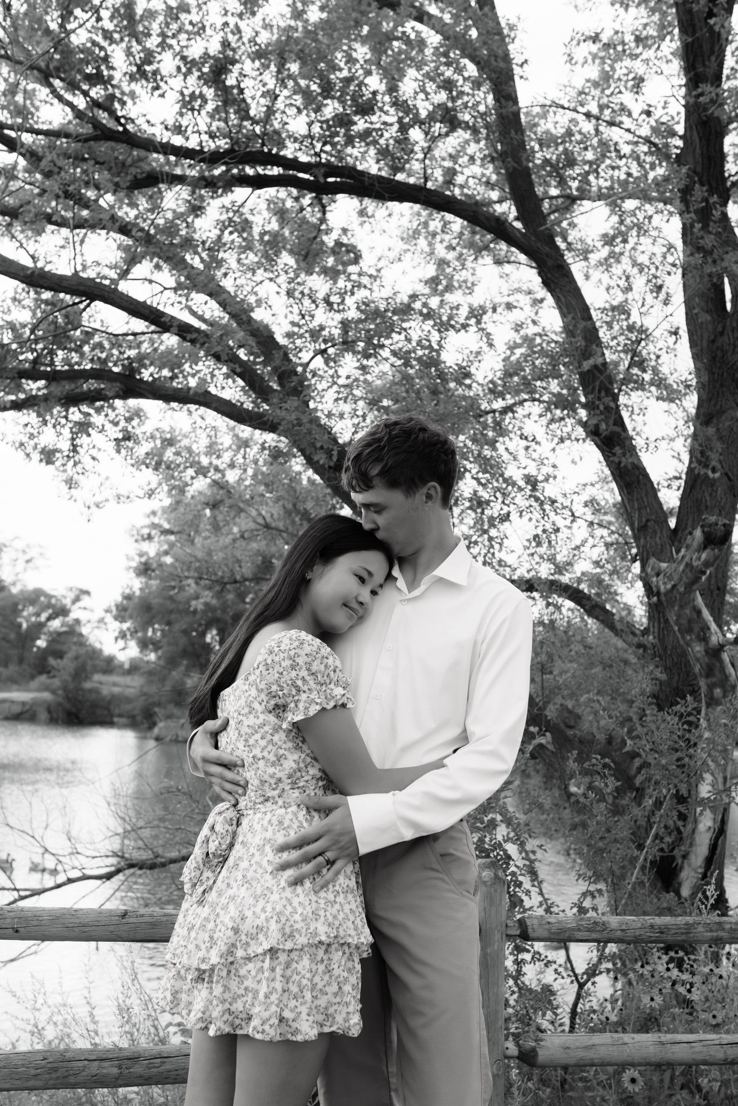 Black and white photo of a couple embracing tenderly by a lakeside fence under large trees.
