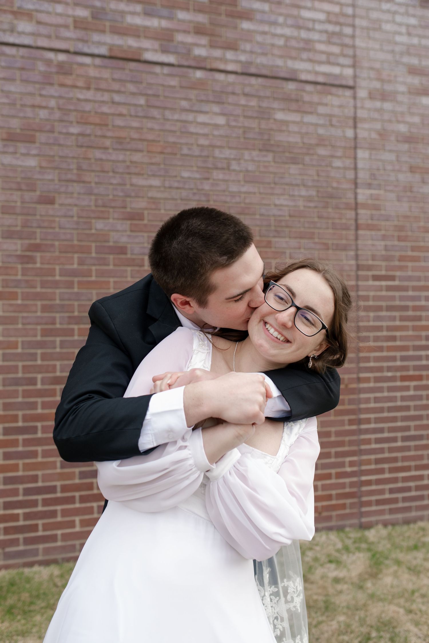 Groom in black suit embracing and kissing bride in white dress on the cheek outdoors by brick wall.