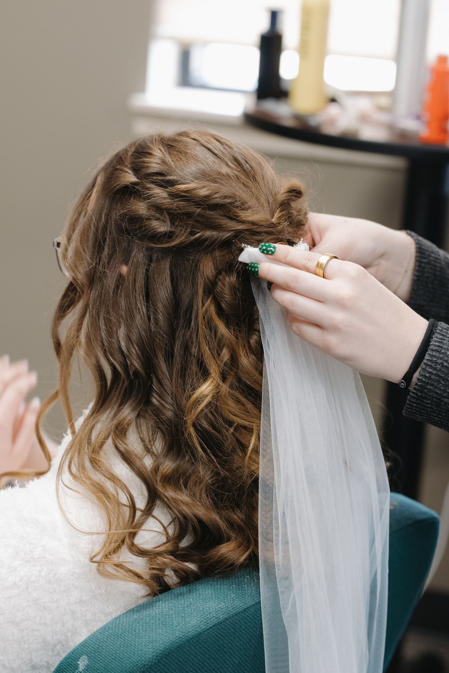 Hairstylist attaching a bridal veil to a bride's half-up curly hairstyle with a braided detail in a salon.