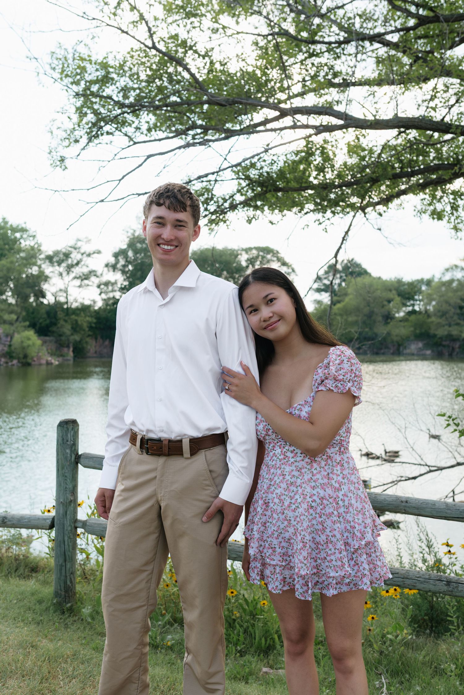 Smiling couple posing by a lakeside fence, man in white shirt and khakis, woman in floral dress.