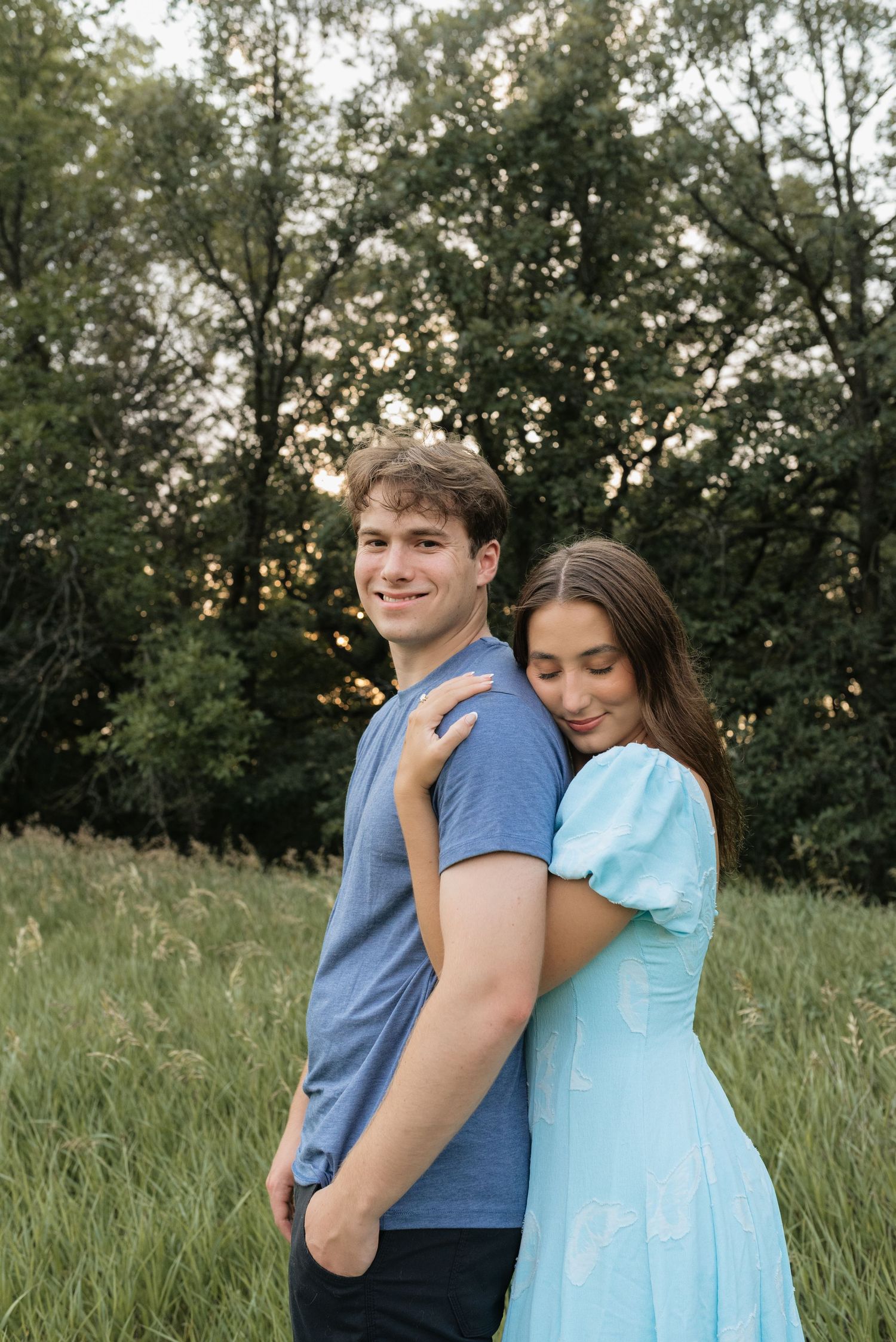A young couple embraces in a lush green field, woman in blue dress hugging man in gray tee, trees in background.