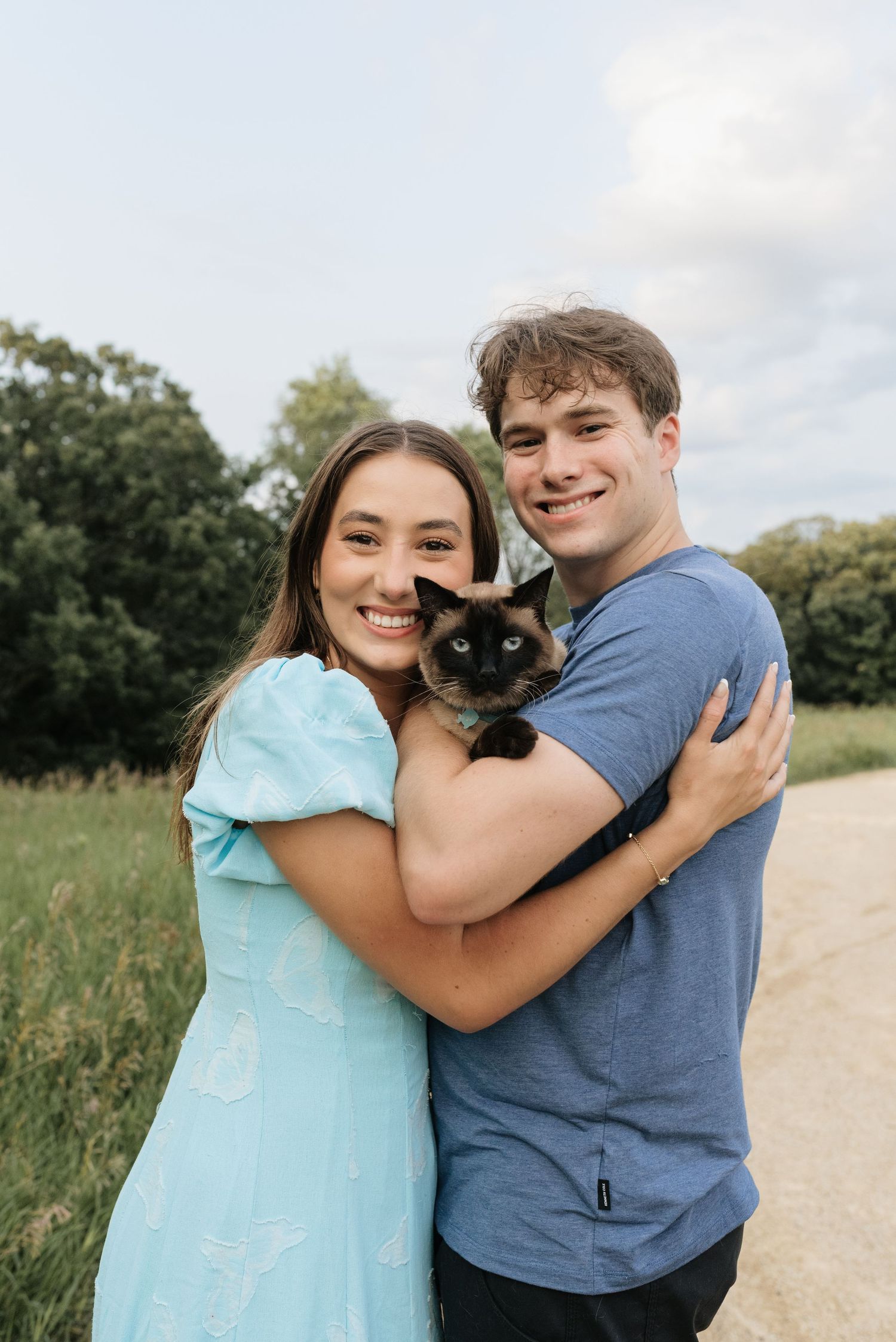 Smiling young couple holding a Siamese cat outdoors on a path surrounded by green trees and grass.