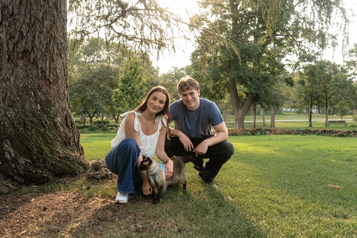 A young couple crouches by a large tree on green grass, posing with a small dog in a sunny park setting.