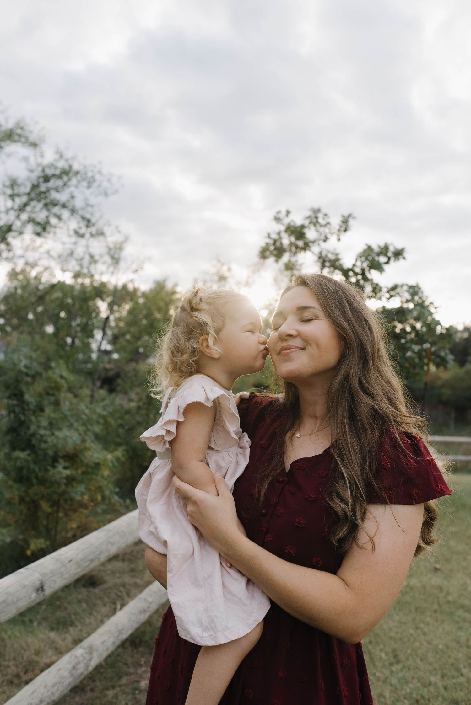 Young mother in a burgundy dress holding her toddler daughter who gives her a sweet kiss outdoors at sunset.