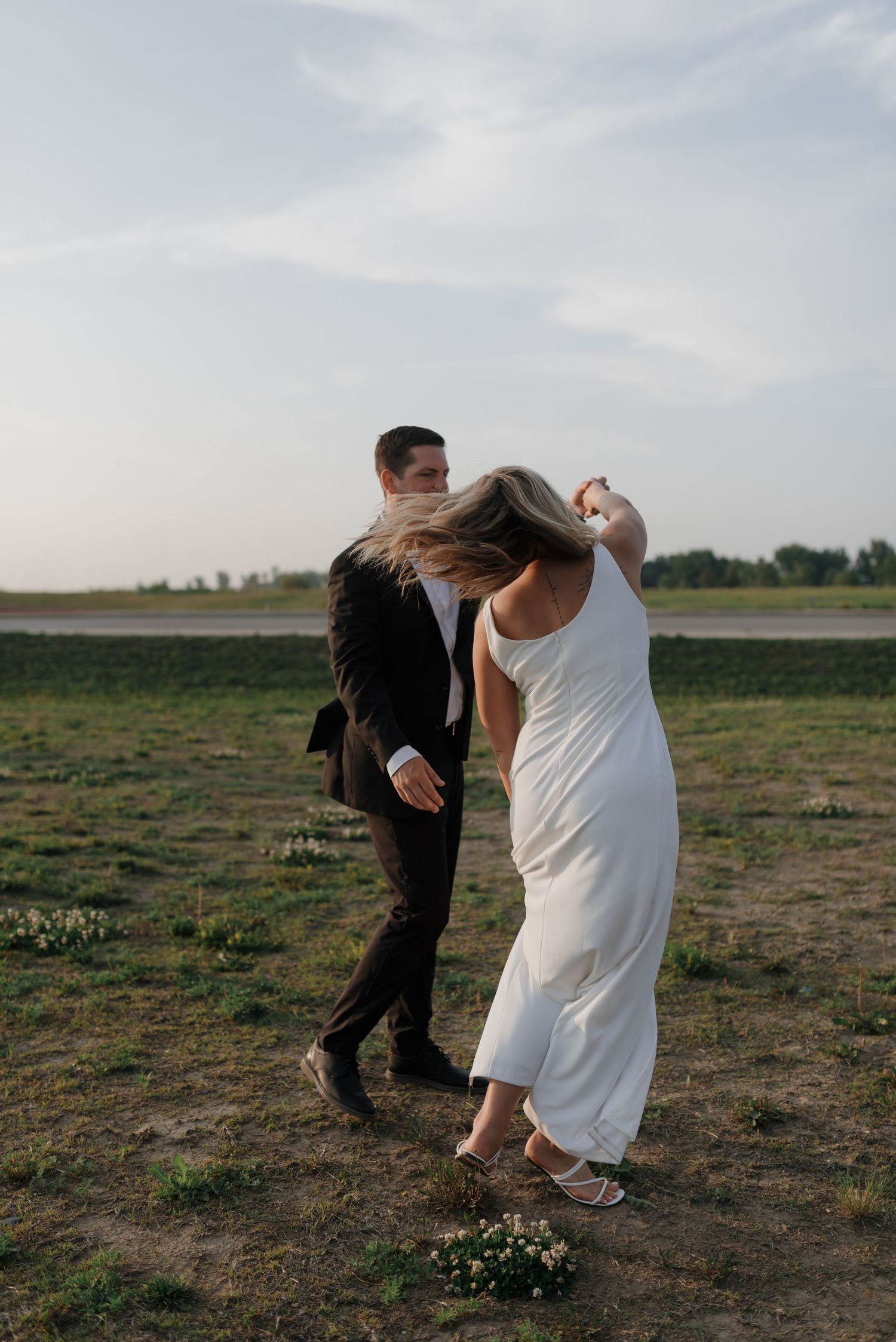 Groom spins bride in white dress outdoors in field during romantic wedding photo shoot.