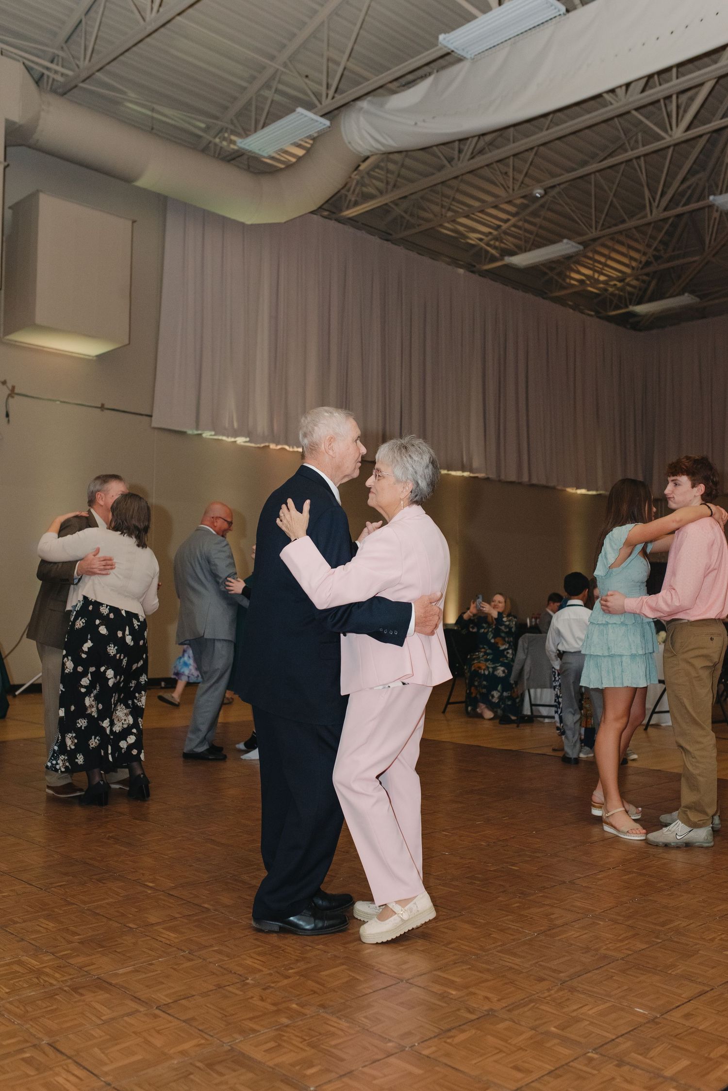 Elderly couple and other guests dancing together on wooden floor at wedding reception.