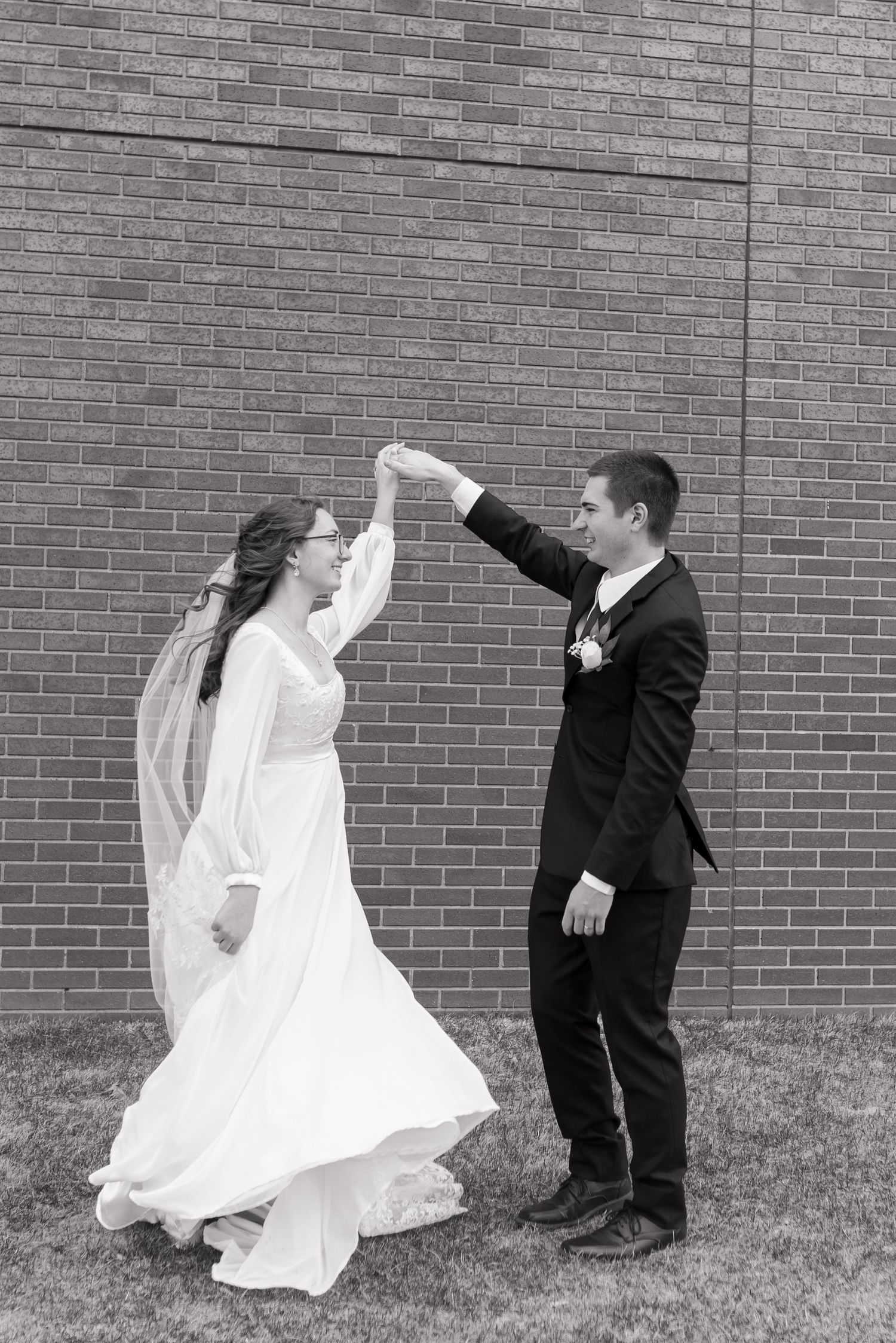 Bride and groom dance outdoors in black and white photo against brick wall in wedding attire.