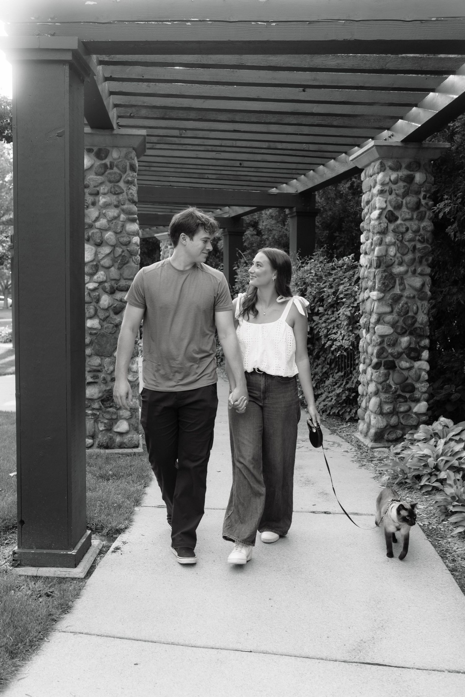 Black and white photo of a couple walking their dog under a pergola, smiling at each other on a sunny day.