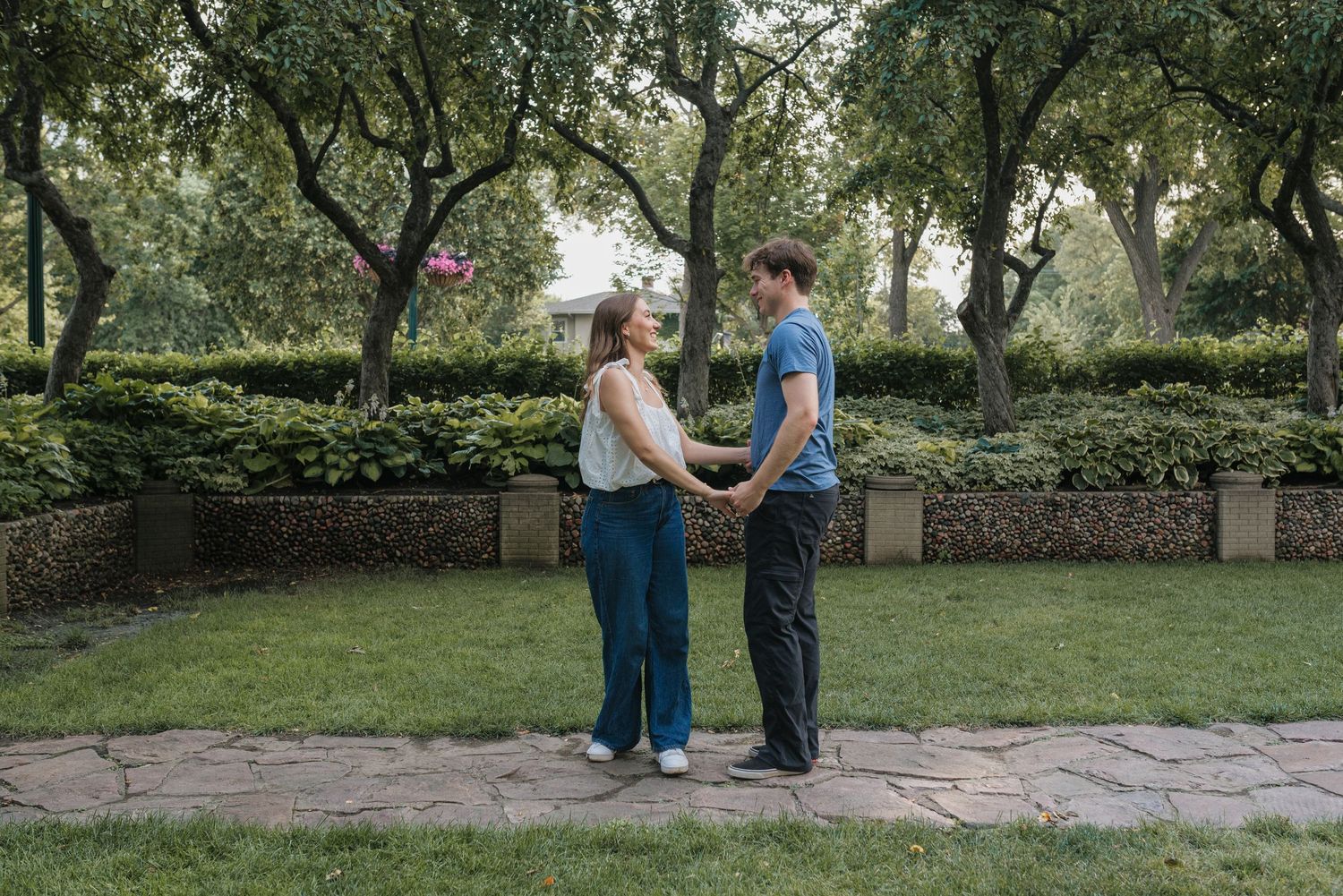 A couple holds hands facing each other in a lush green park surrounded by trees and hedges.