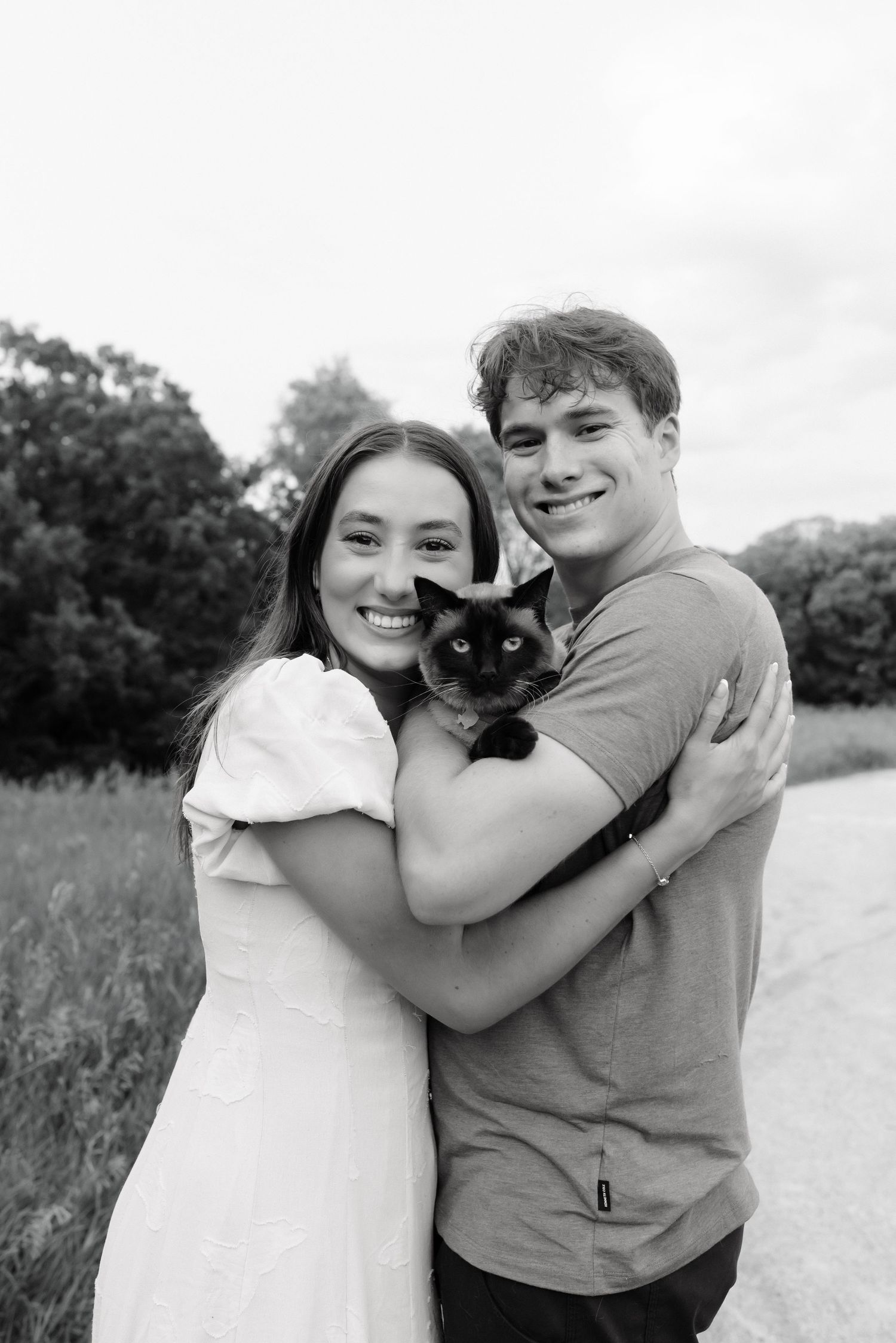 Black and white photo of a smiling young couple outdoors holding a Siamese cat, surrounded by trees.