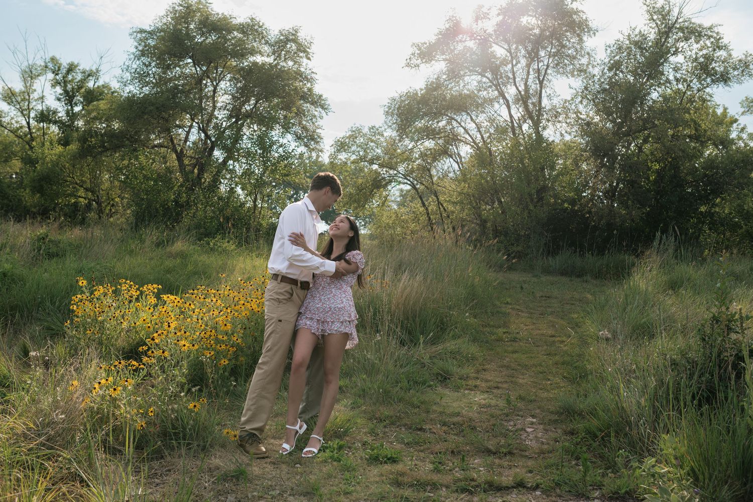 A couple embraces on a sunlit path surrounded by wildflowers and lush greenery.