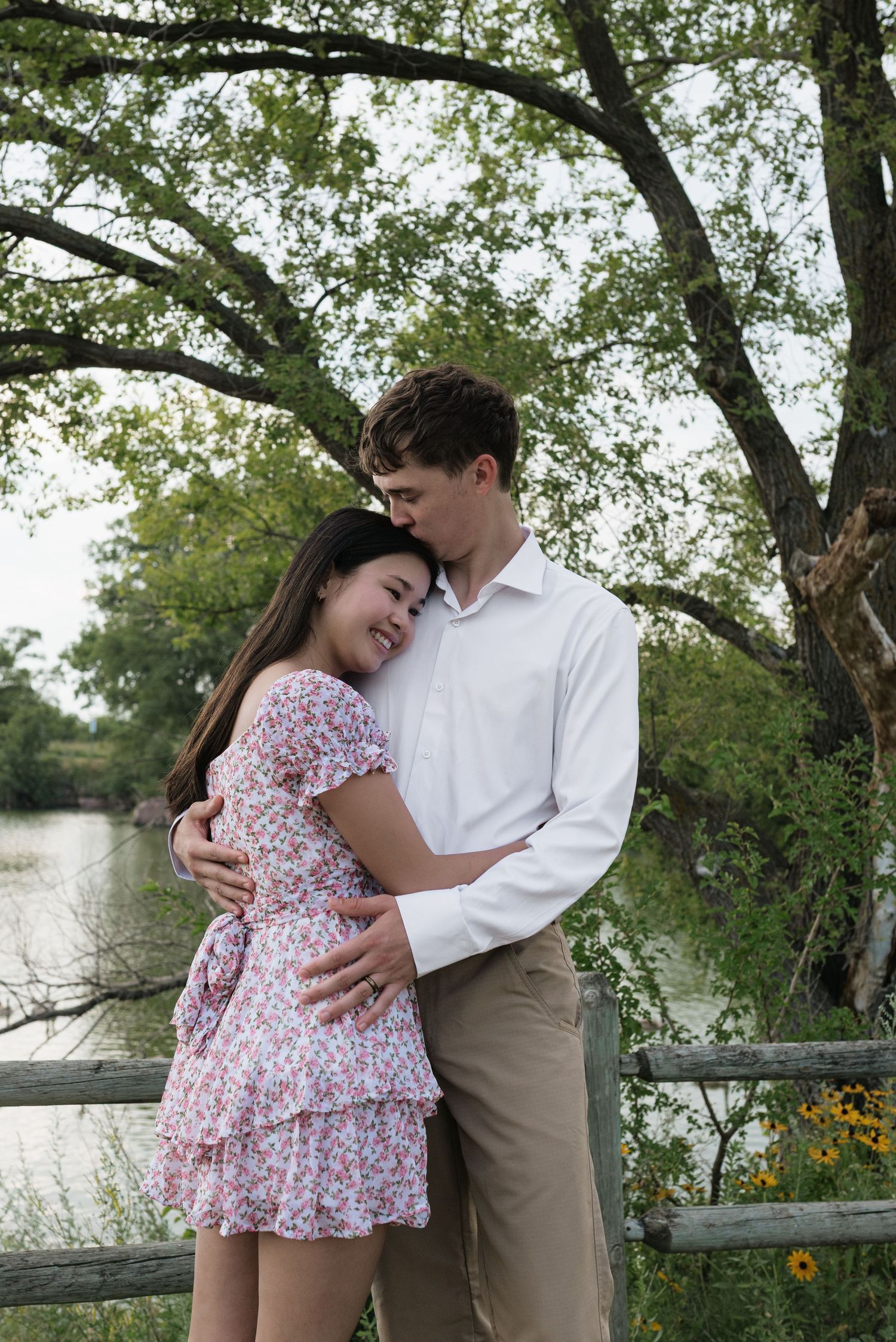 Couple embracing near a wooden fence by a lake, woman in floral dress leaning into man in white shirt.