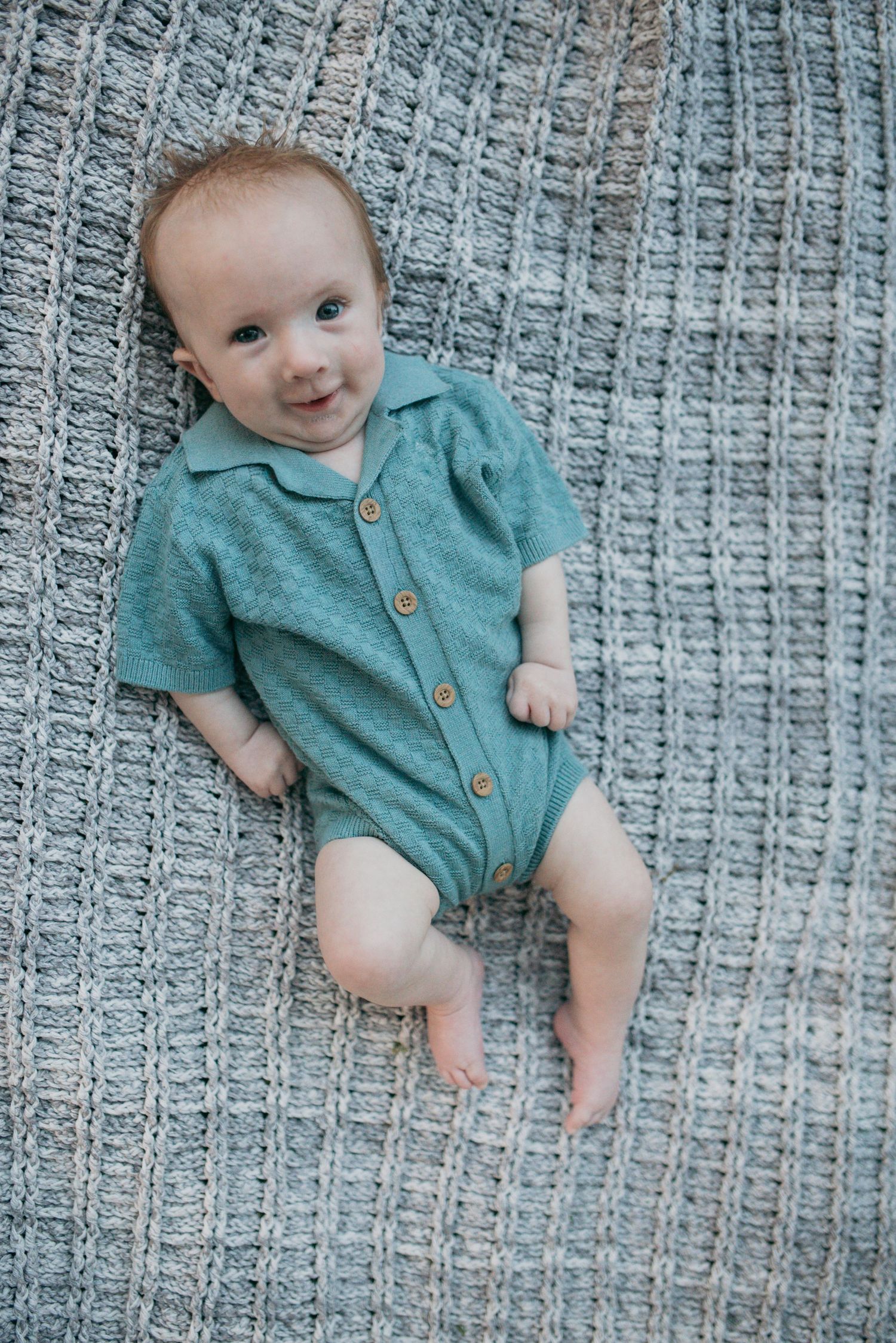 A happy baby in a teal button-front onesie lying on a gray knit blanket, looking up.