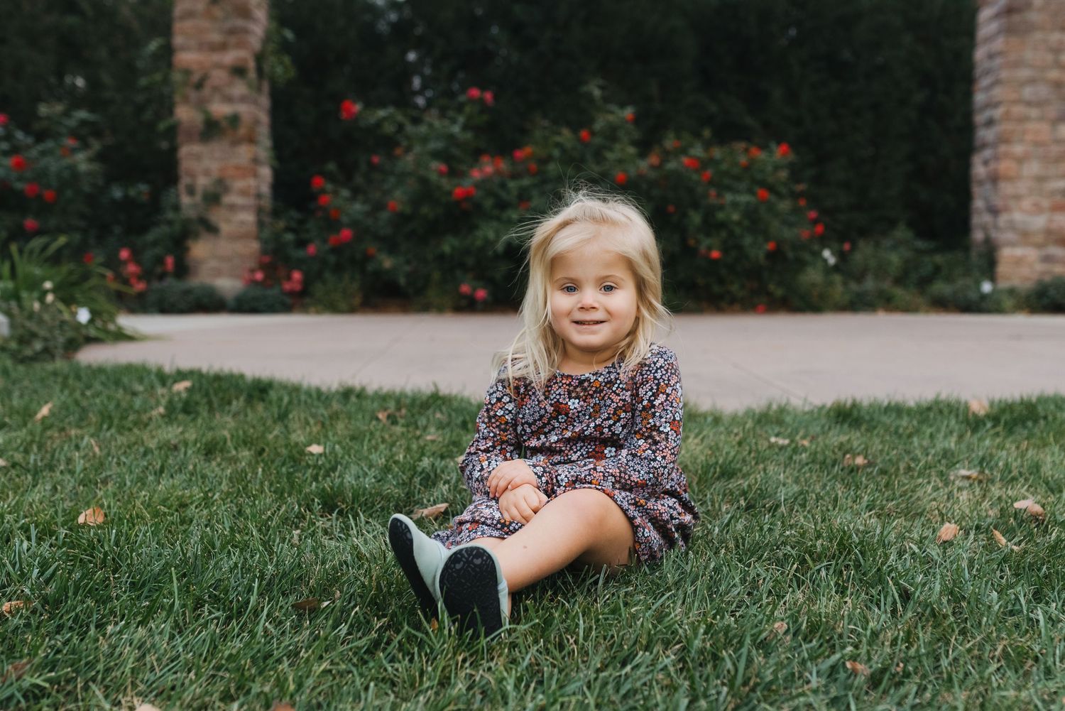 Sweet toddler girl in a floral dress sitting on lush green grass in a beautiful garden setting.