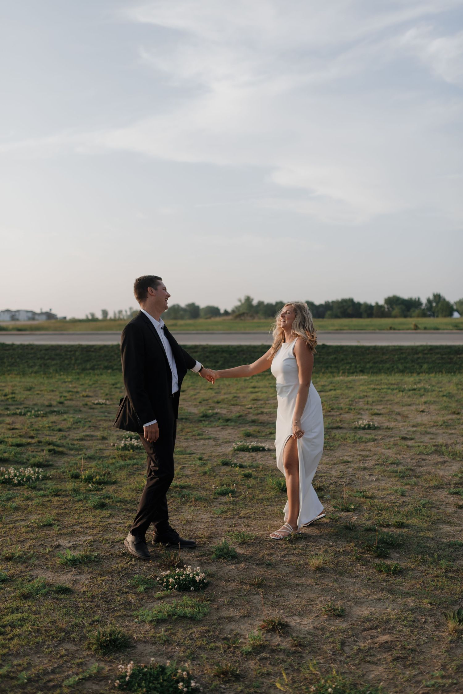 Bride and groom hold hands walking through green field during golden hour wedding photo session.