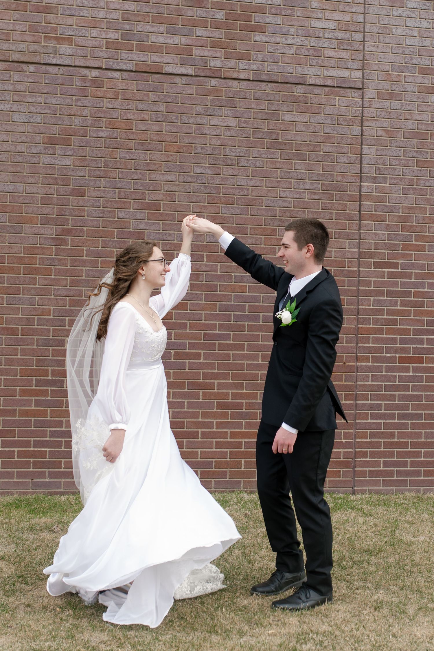 Bride and groom dancing outdoors near brick wall, groom twirling bride in white wedding dress and veil.