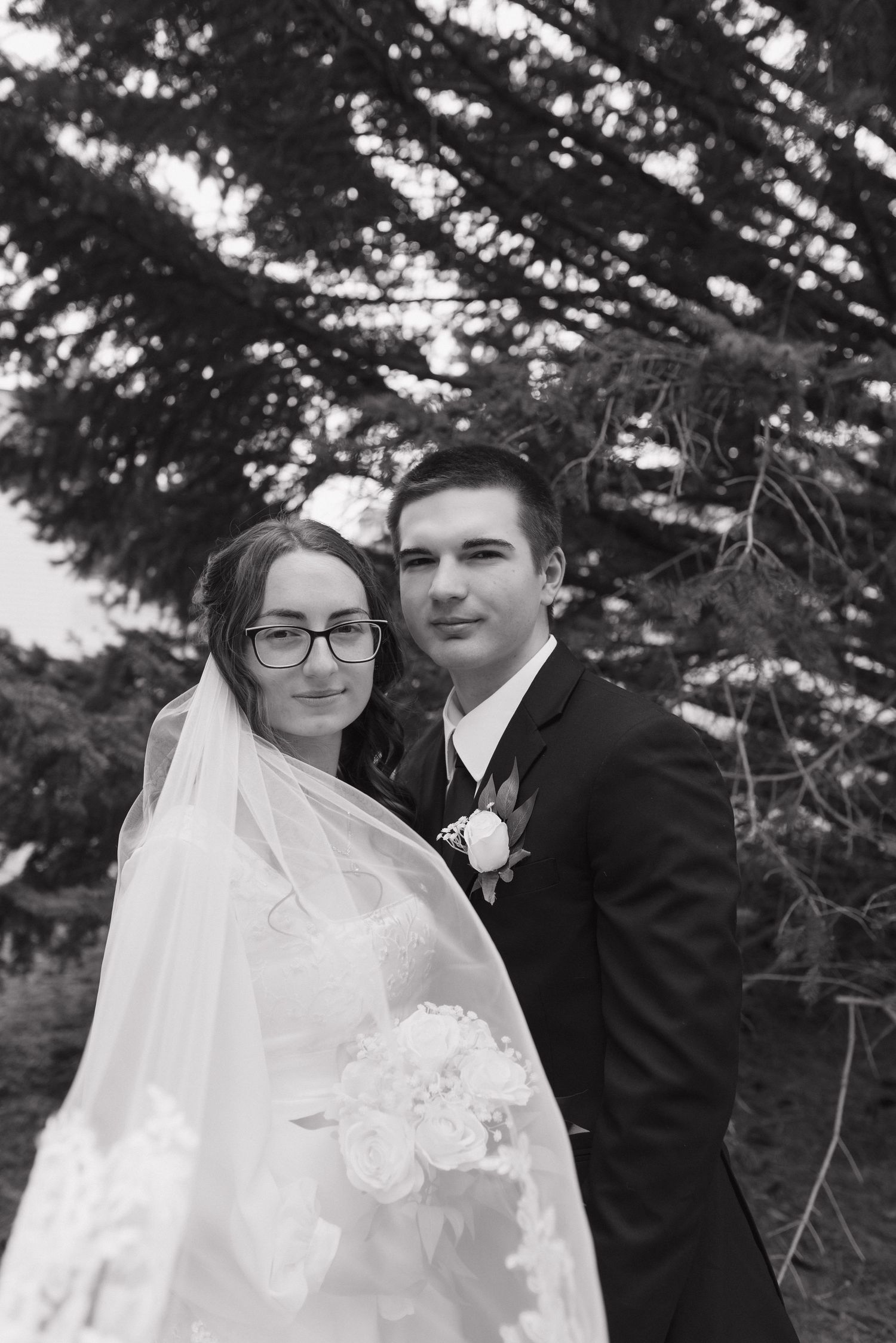 Black and white wedding portrait of bride with veil and bouquet and groom in suit posing before evergreen trees.