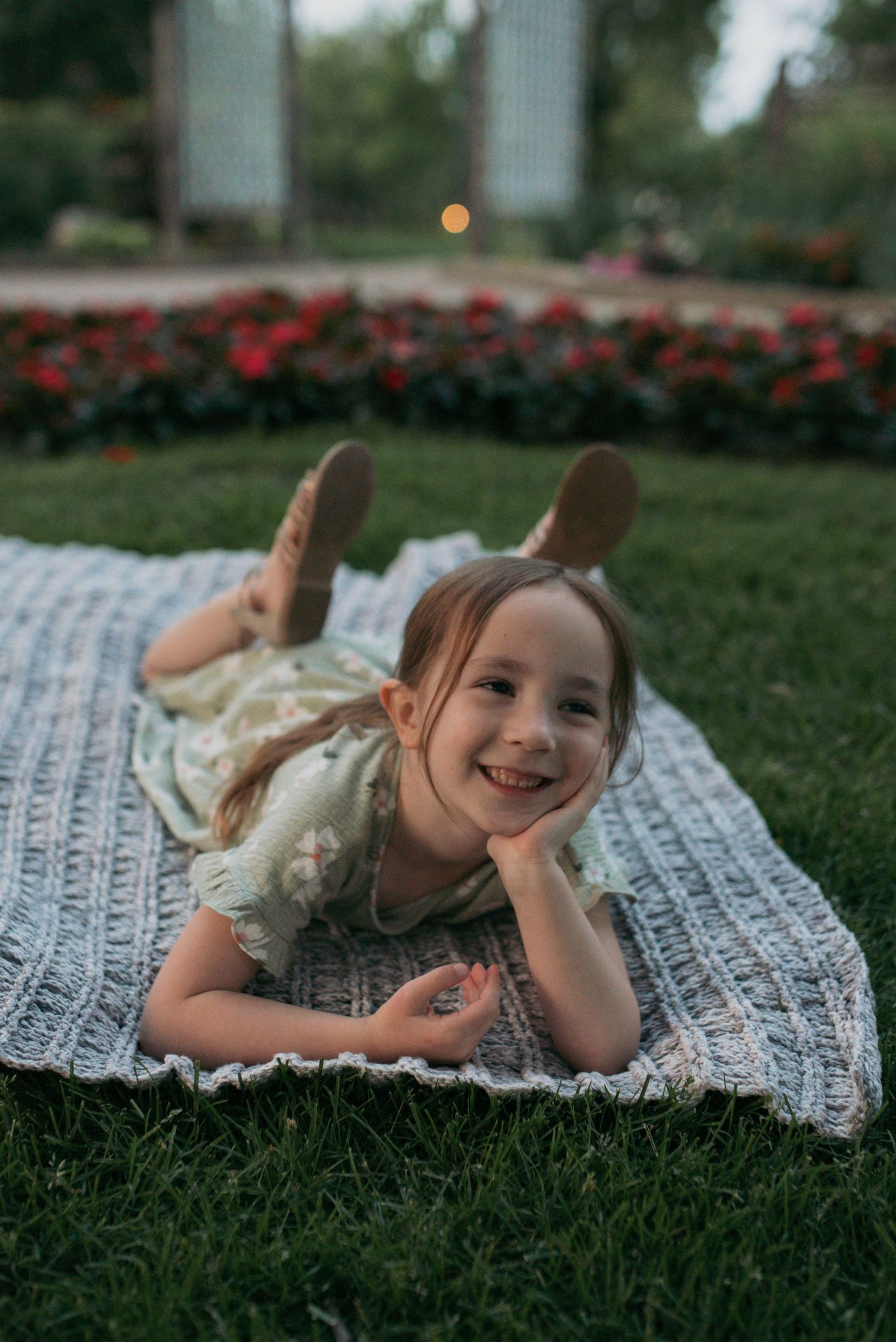 A happy little girl lying on a crochet blanket outdoors, smiling with red flowers behind her.