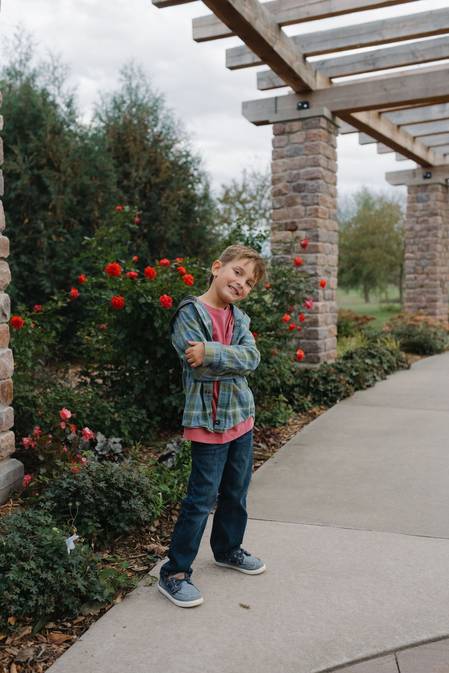 Young boy in plaid jacket and jeans standing with arms crossed near rose bushes under a pergola.