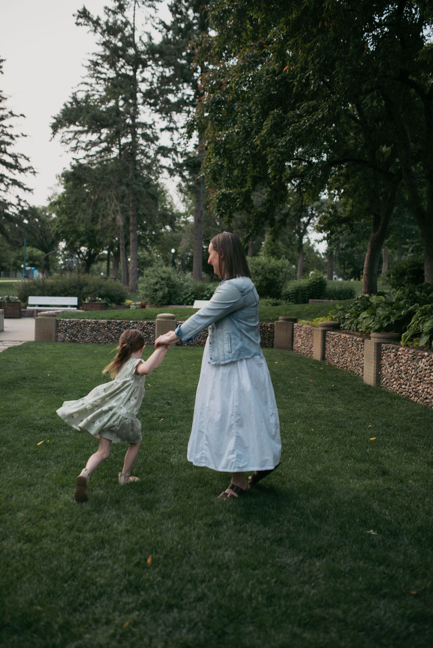 Mother in denim jacket spins young daughter in floral dress on park lawn.